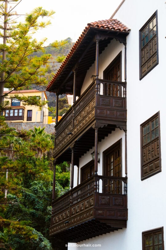 White building with two dark wooden balconies, detailed railings, and large windows, set against greenery and a hillside in the backgroundâ€”typical of Garachico Tenerifeâ€™s charming architecture.