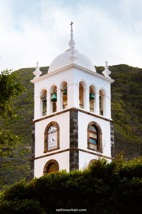 A white bell tower with a clock and green bells stands against a backdrop of green hills and a partly cloudy sky in Garachico, Tenerife.