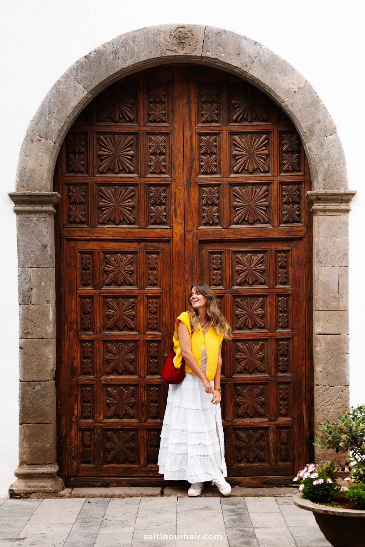 A woman in a white dress and yellow top stands in front of a large, ornately carved wooden door set in a stone archway in Garachico, Tenerife.