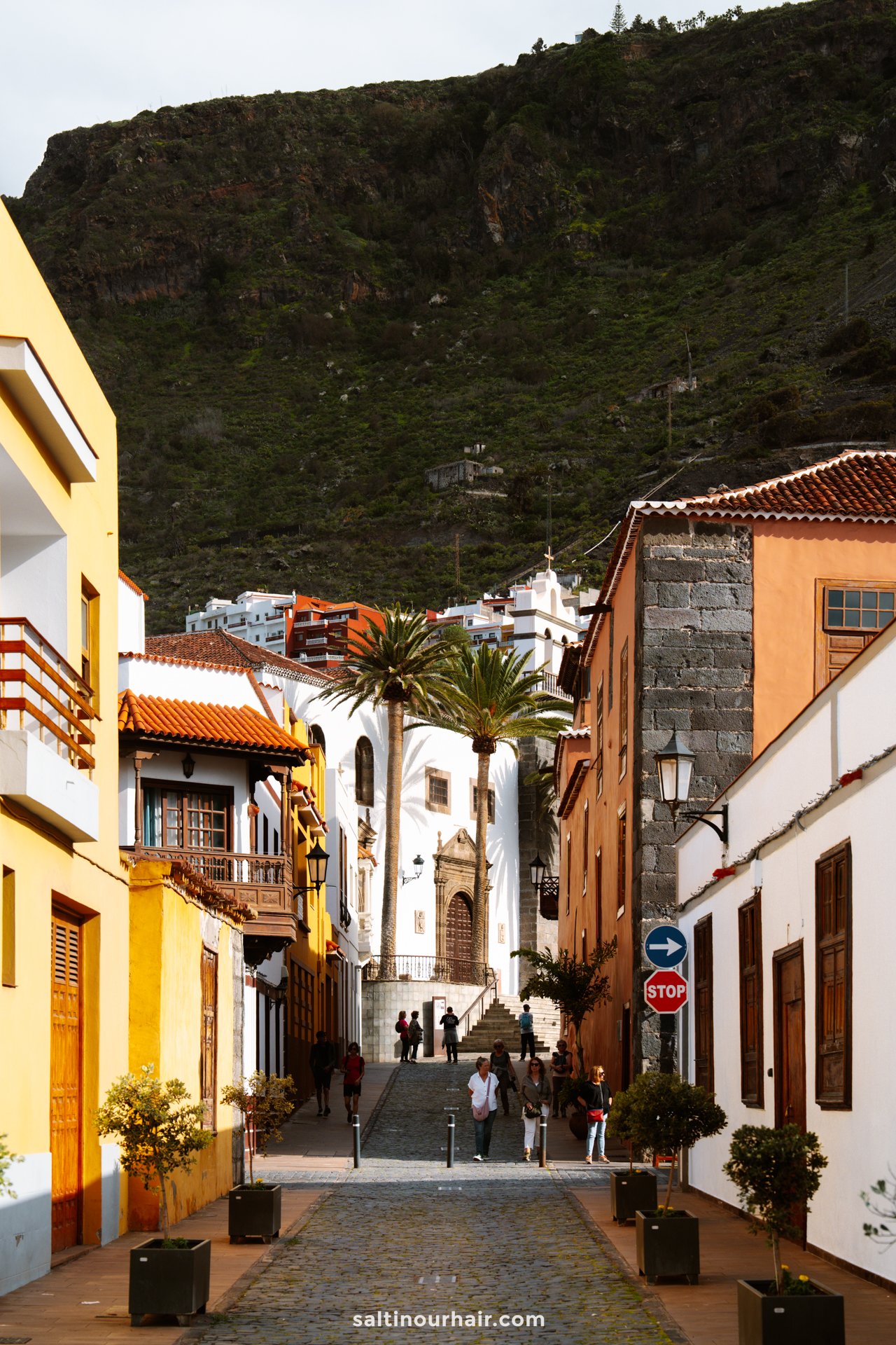 A cobblestone street with colorful buildings leads to a white church in Garachico, Tenerife, with people walking and a green mountain in the background.