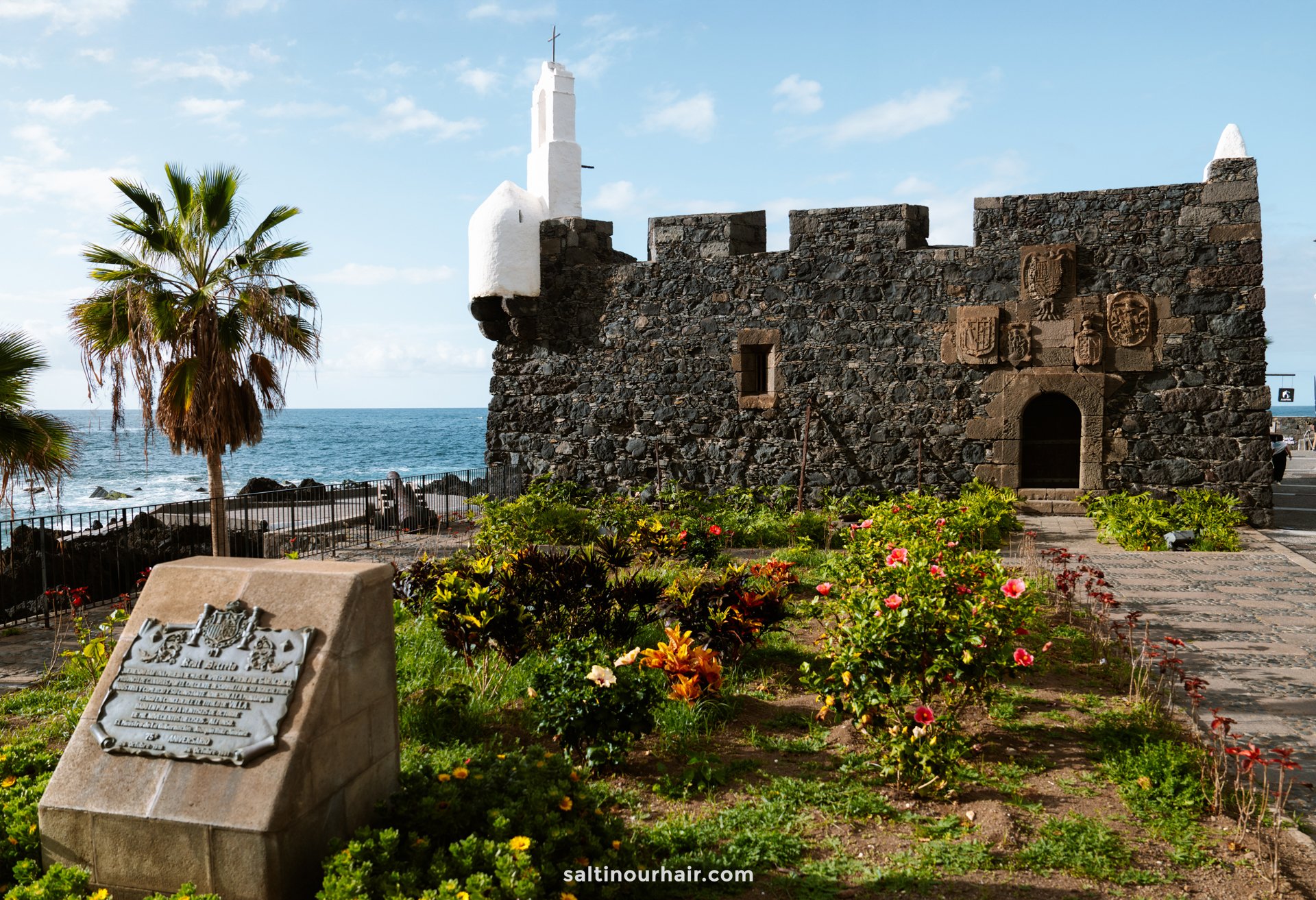 A historic stone fortress with a small white tower stands near the ocean in Garachico, Tenerife, surrounded by gardens, palm trees, and a commemorative plaque in the foreground.