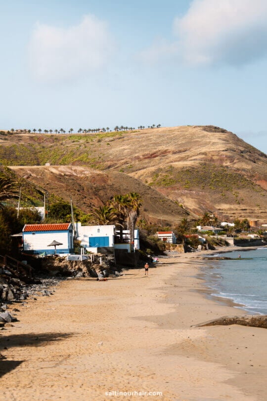 A sandy beach with gentle waves, a few small white houses, palm trees, and a dry, hilly landscape in the background under partly cloudy skiesâ€”perfect for relaxing or discovering things to do in Porto Santo.