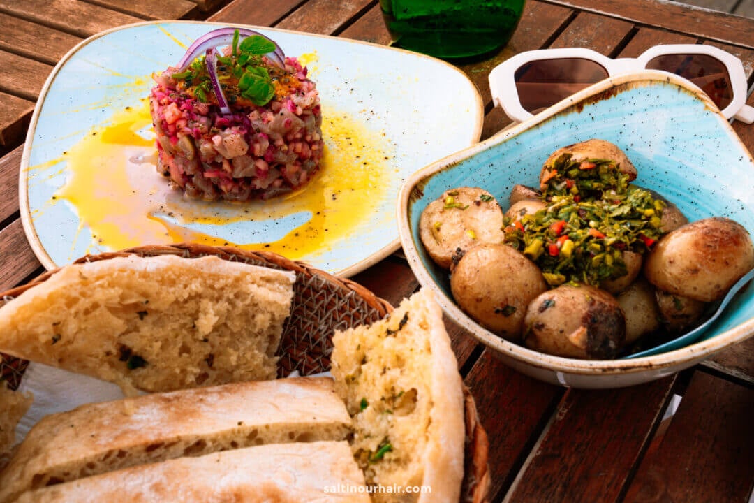 A plate of bread slices, a bowl of seasoned baby potatoes, and a dish of tuna tartare with red onions and herbs await on a wooden tableâ€”perfect for sampling local flavors after exploring things to do in Porto Santo.