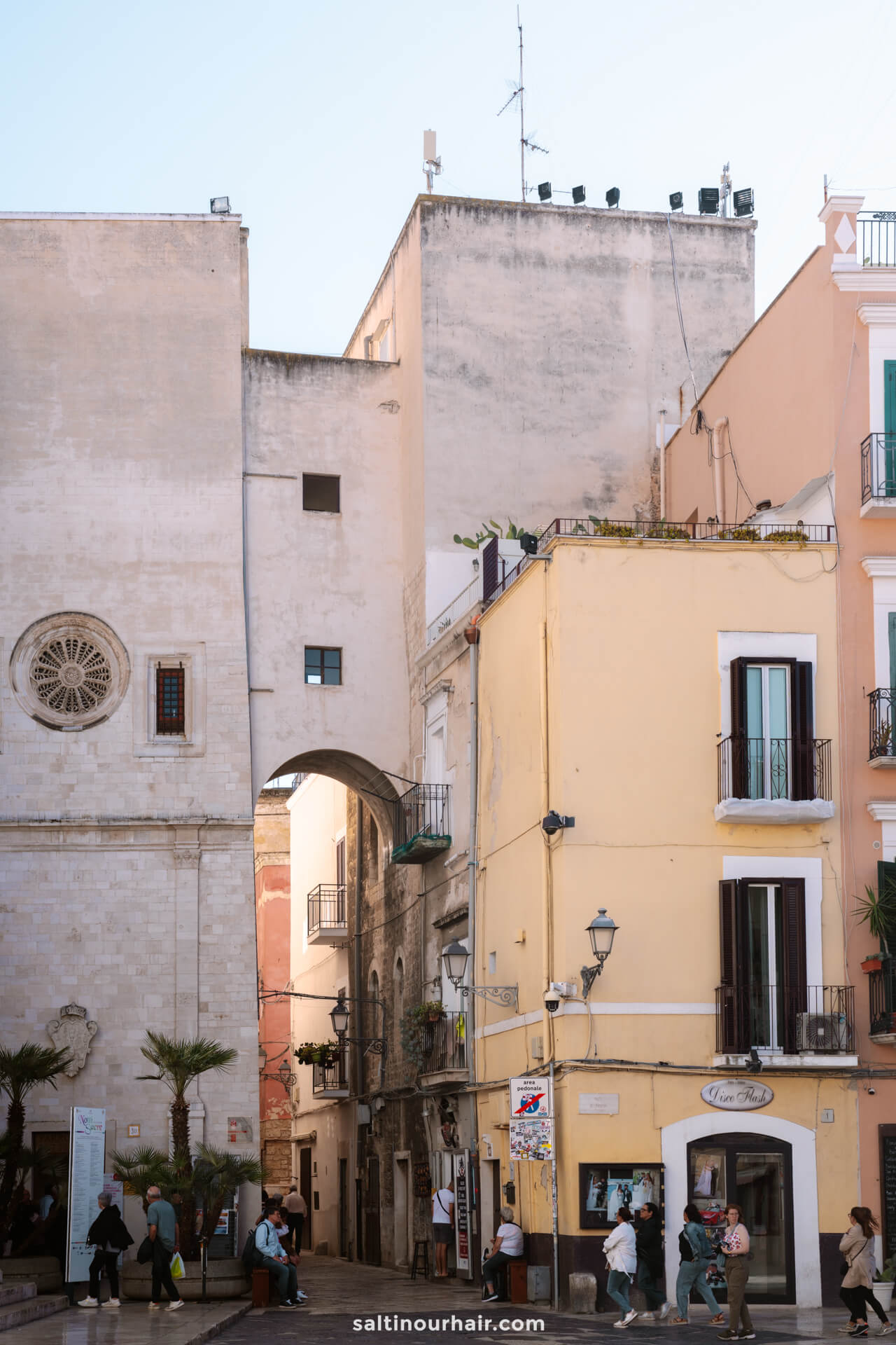 A narrow archway connects old stone buildings with pastel facades in a town square in bari puglia italy, where balconies overlook a lively scene