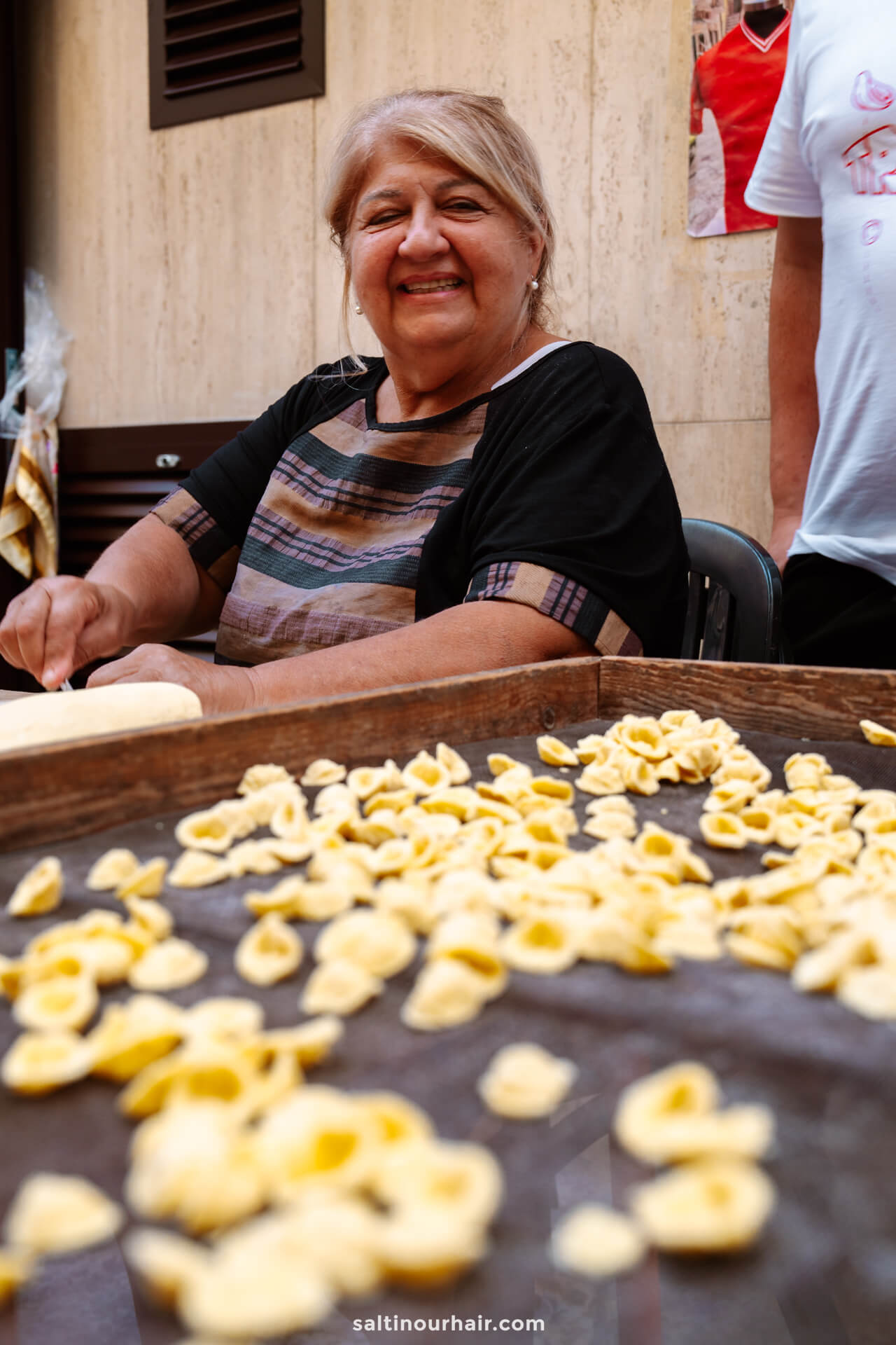 An older woman sits at a table smiling while making fresh pasta, with handmade pasta pieces spread out on a tray in front of her in Bari, Italy