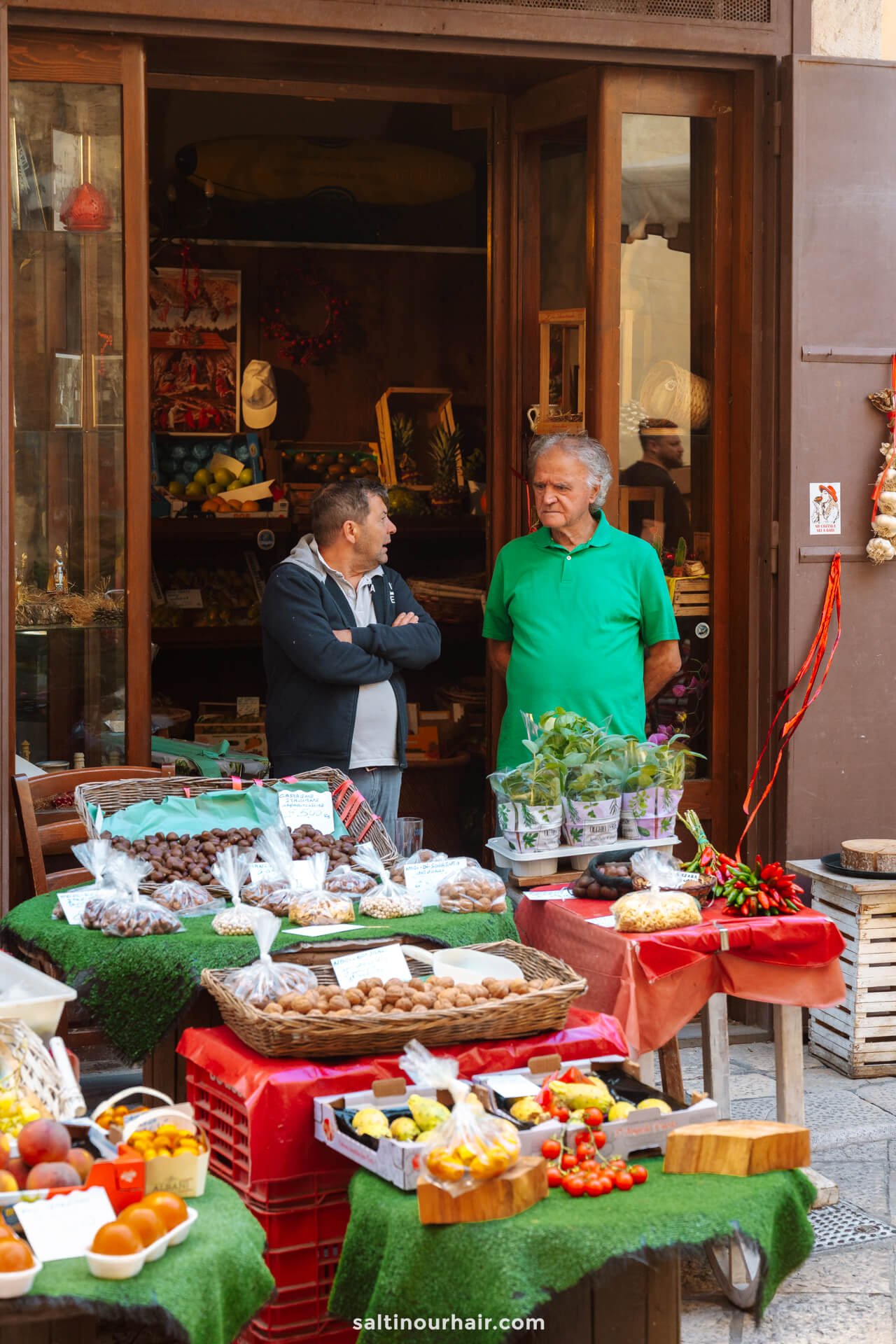 Two men stand and talk in front of a small shop in Bari, italy selling fresh produce, baked goods, and packaged foods displayed outside