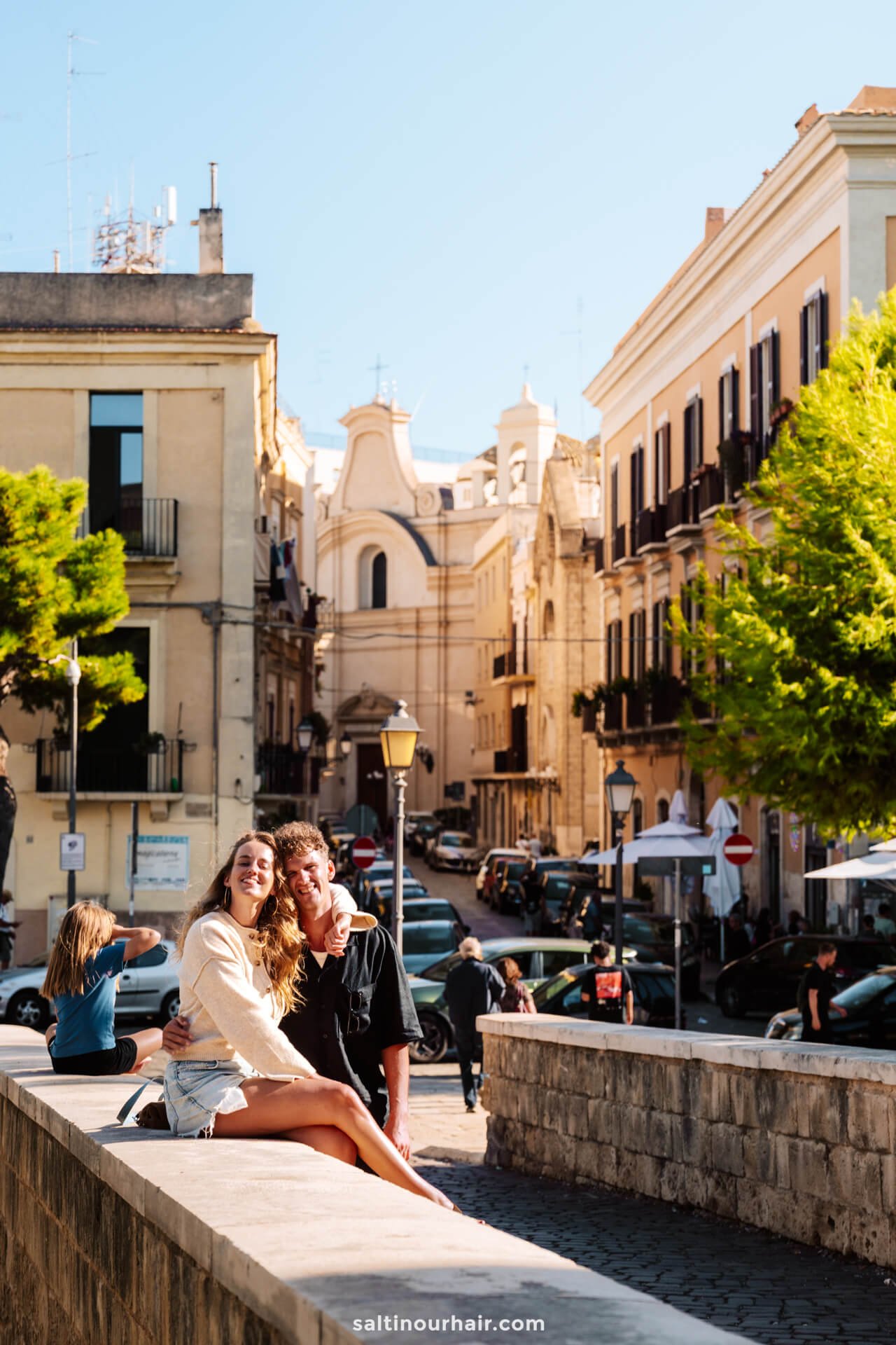 A young couple sits on a stone ledge in a sunlit Bari, Italy, surrounded by historic buildings and parked cars