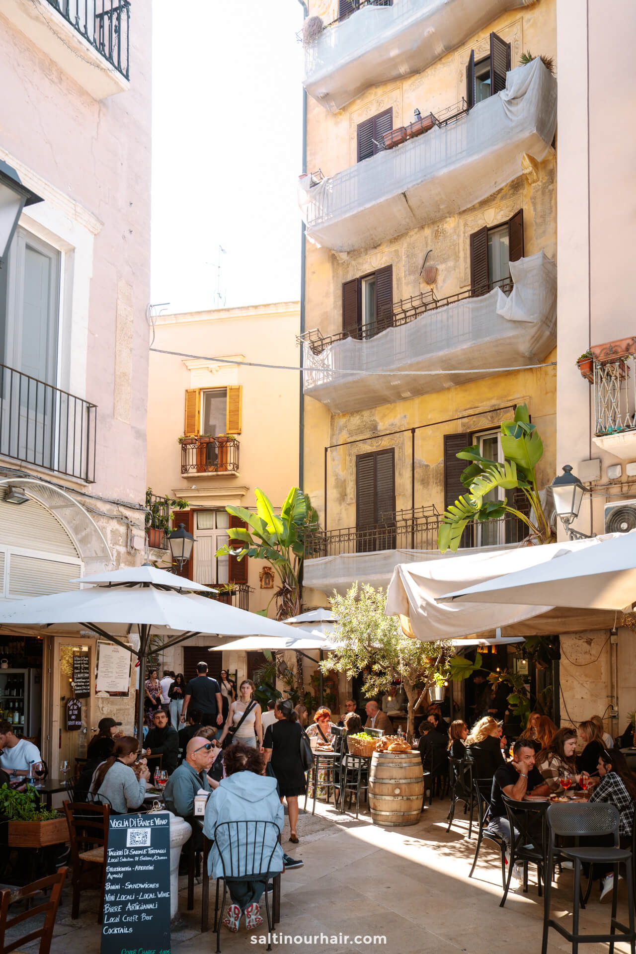 Outdoor café with people dining under large umbrellas in a narrow courtyard, surrounded by tall, weathered apartment buildings with balconies and plants—a charming stop among the many things to do in Bari puglia italy