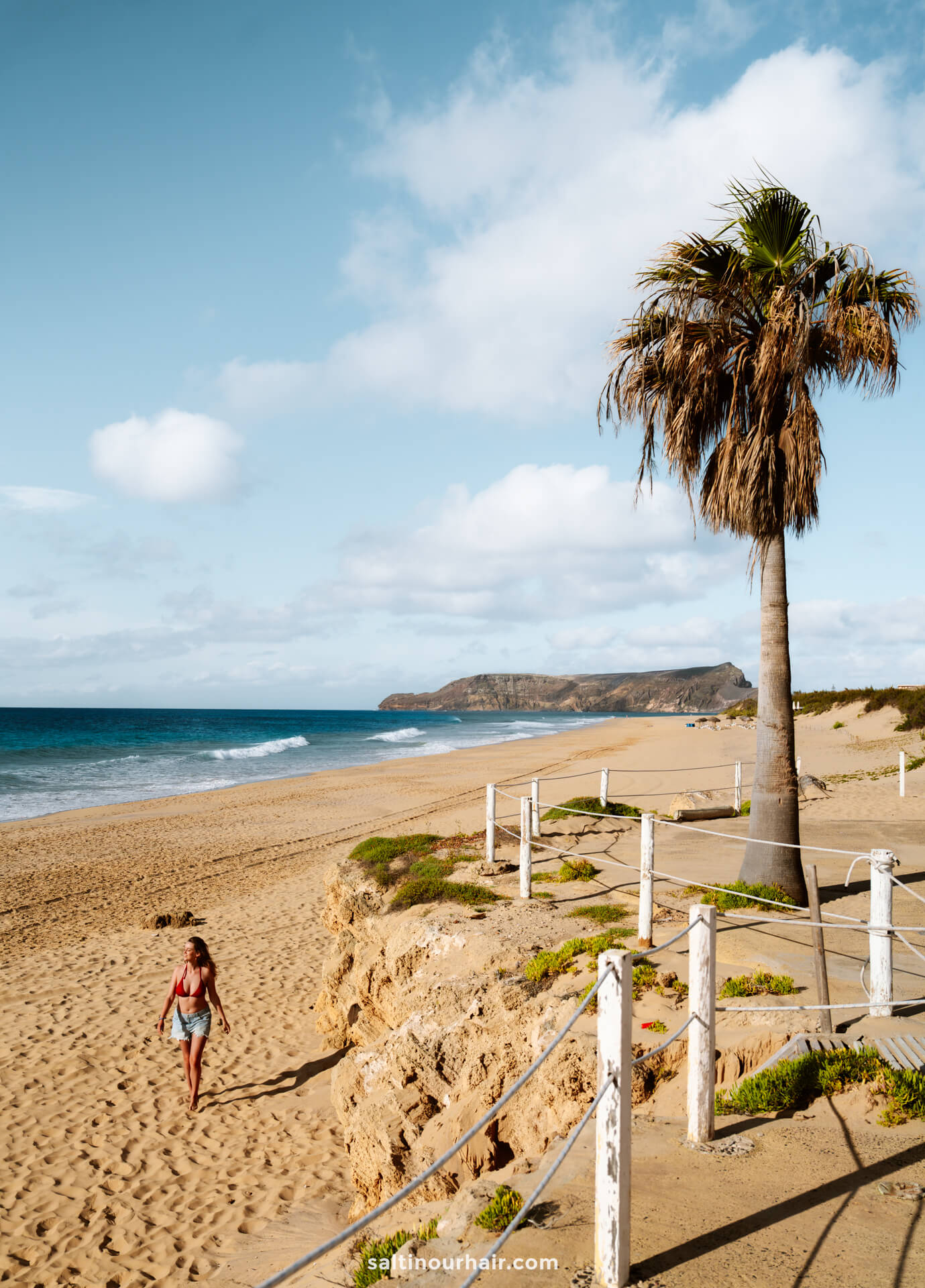 A woman walks along a sandy Porto Santo beach next to a palm tree, with the ocean and distant hills under a partly cloudy skyâ€”one of the many relaxing things to do in Porto Santo.