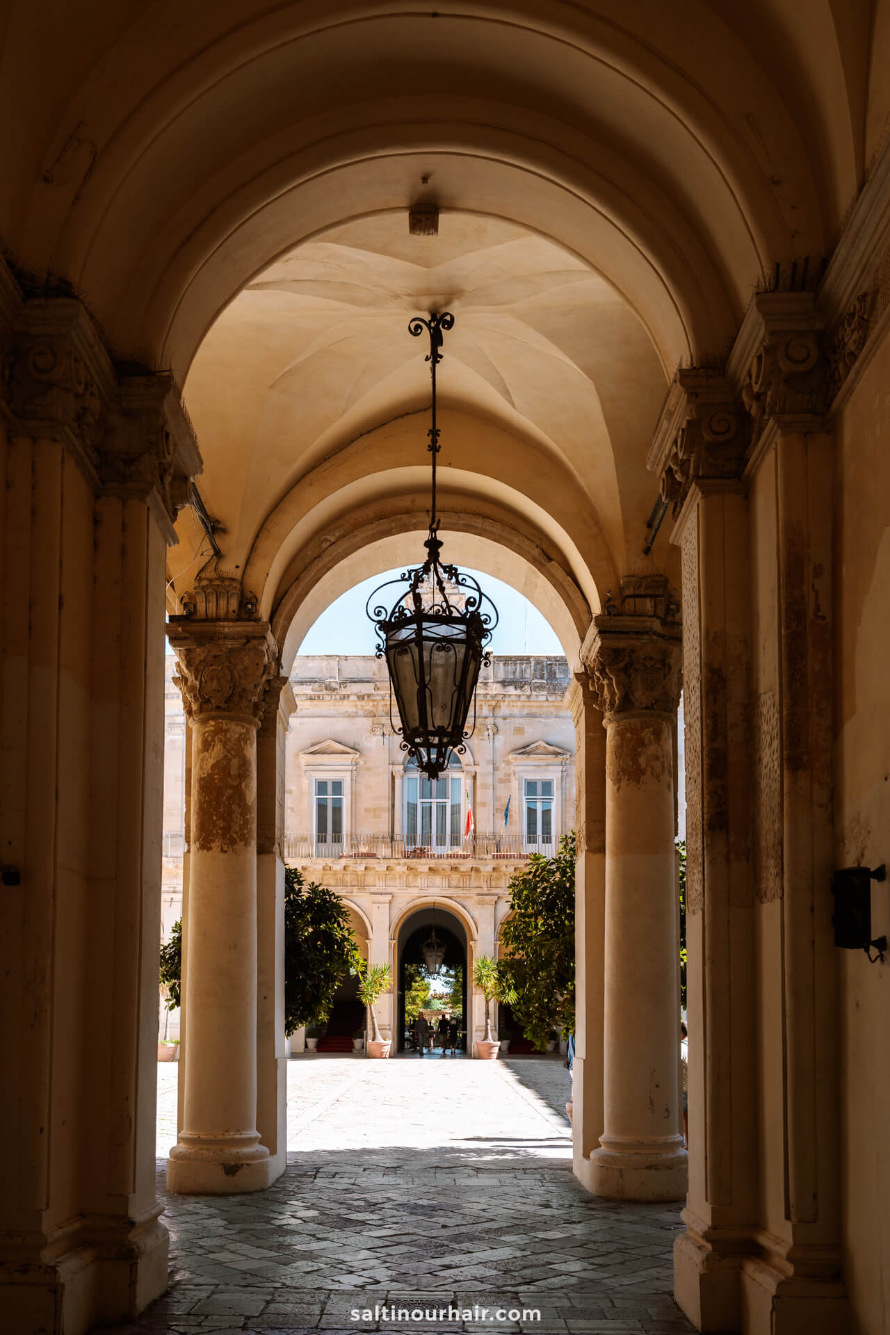 An archway with a hanging lantern frames the view of a sunlit courtyard in Lecce, Italy