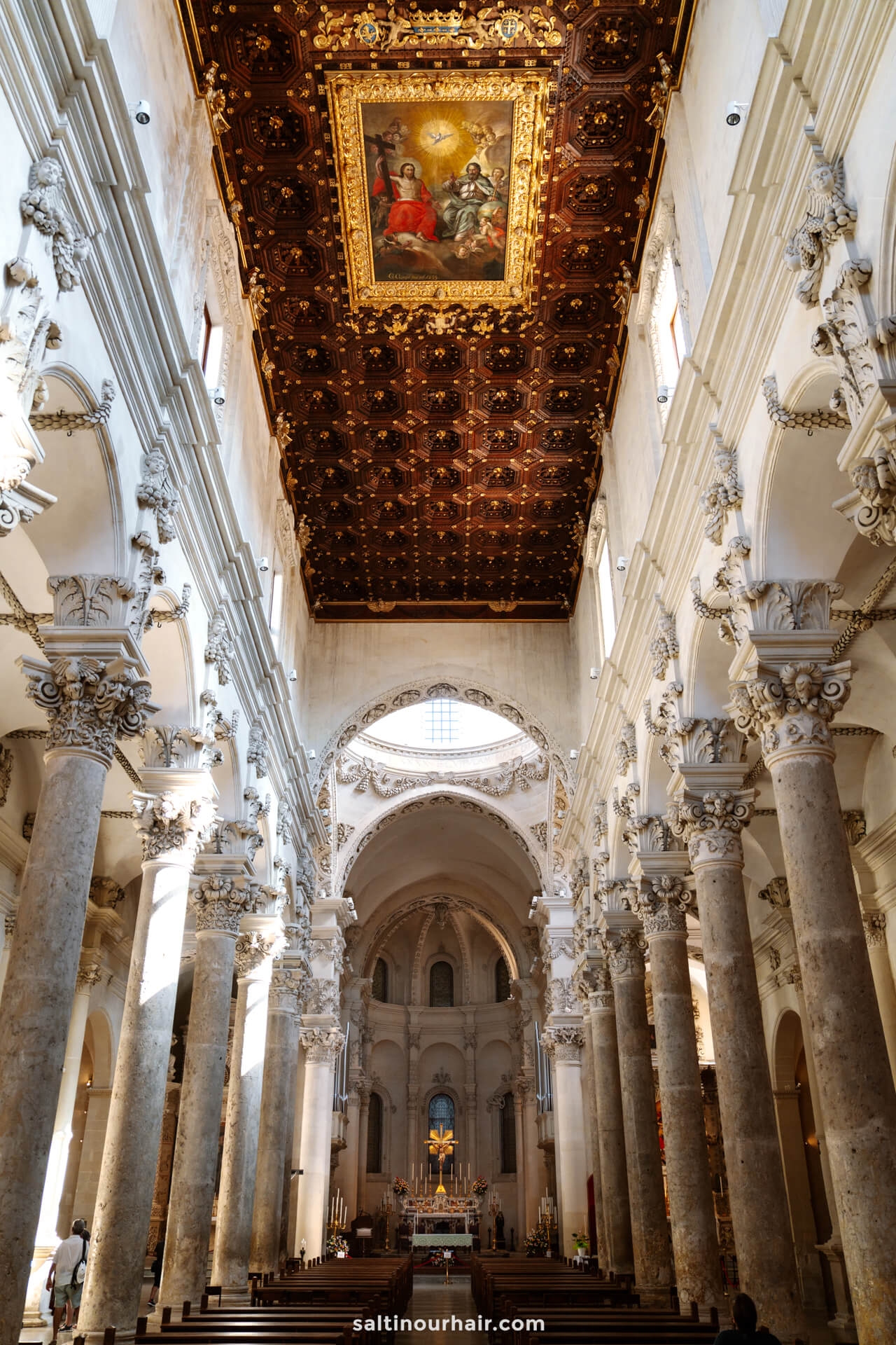 Interior view of Basilica di Santa Croce in Lecce, Italy, with marble columns and an ornate wooden ceiling featuring a large religious painting
