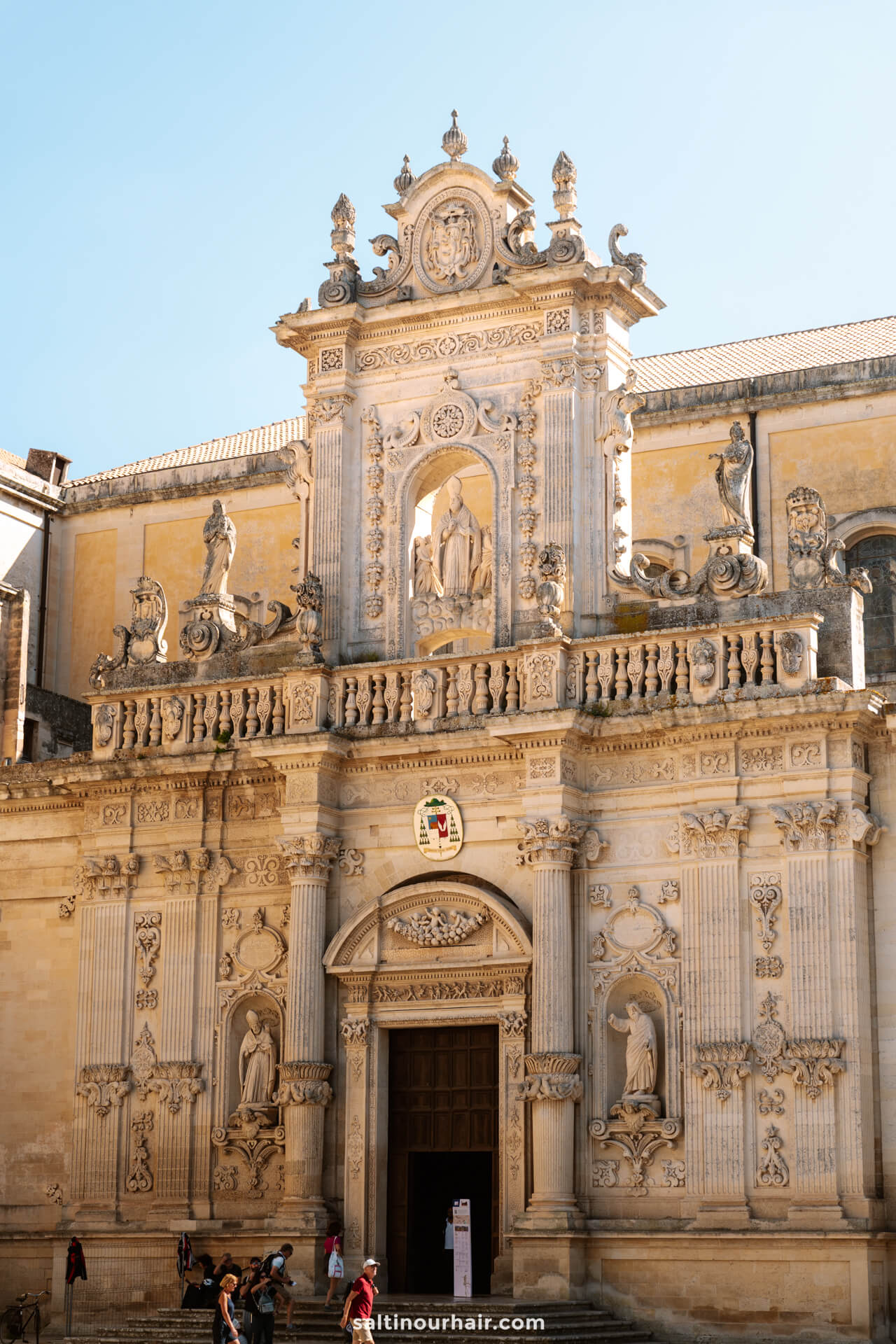 Historic building in Piazza del Duomo in Lecce, Italy, with statues, columns, ornate carvings, and people standing near the entrance