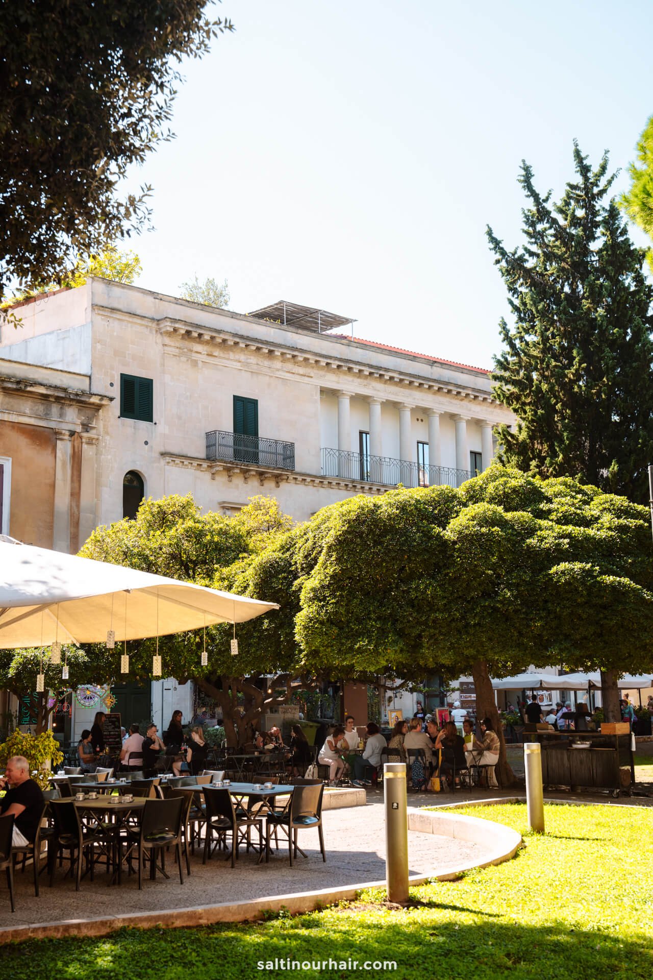 Outdoor café with people sitting at tables under large trees and umbrellas, adjacent to a beige building with green shutters and a balcony on a sunny day in Lecce, Italy.