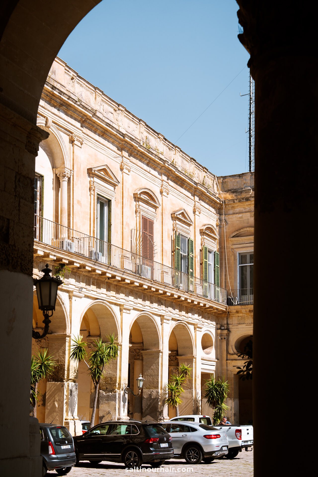View of a historic beige building in Lecce, Italy, featuring arched walkways, balconies, and potted palm trees