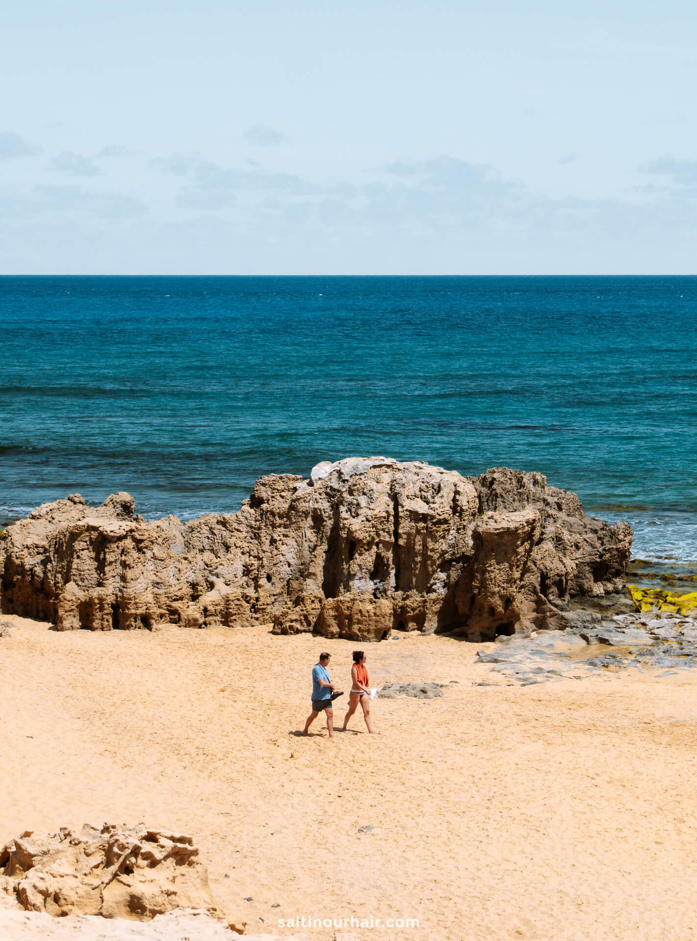 Two people walking on a sandy Porto Santo beach with rocky formations and the ocean in the background under a partly cloudy skyâ€”one of the top things to do in Porto Santo.