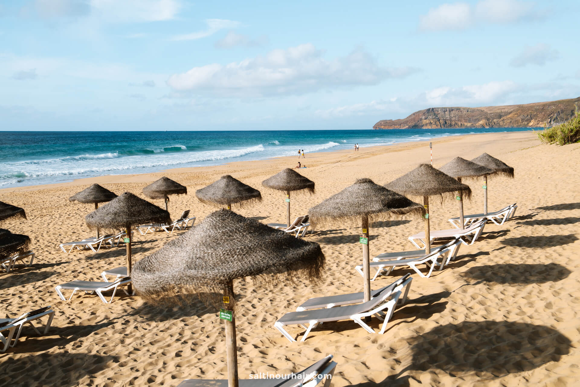 Rows of empty lounge chairs with thatched umbrellas on a sandy Porto Santo beach, overlooking the ocean and distant hills under a partly cloudy skyâ€”a perfect setting when searching for things to do in Porto Santo.