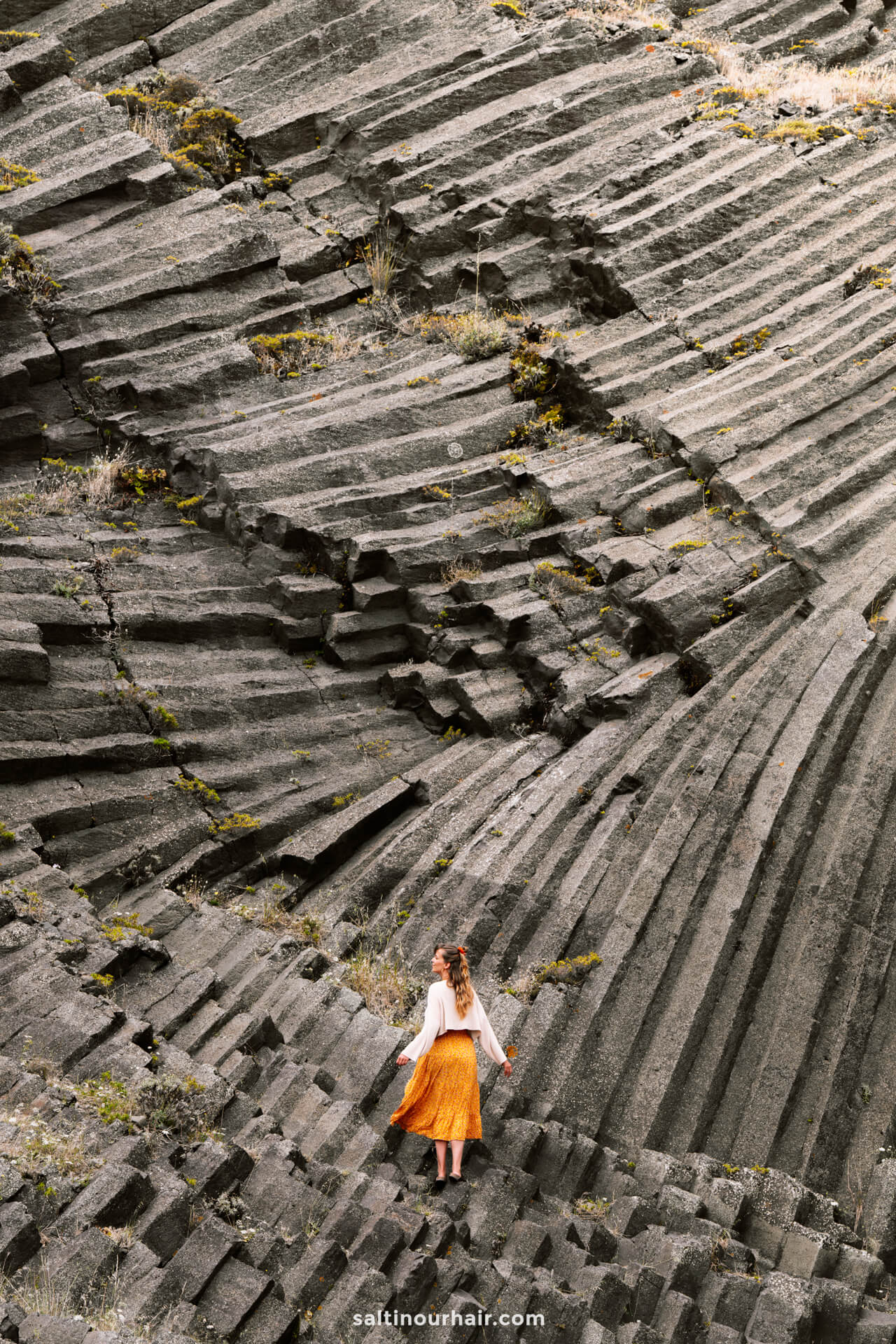A woman in a white top and orange skirt stands on large, layered rock formations with dramatic, angular linesâ€”a striking scene among the unique landscapes of Porto Santo