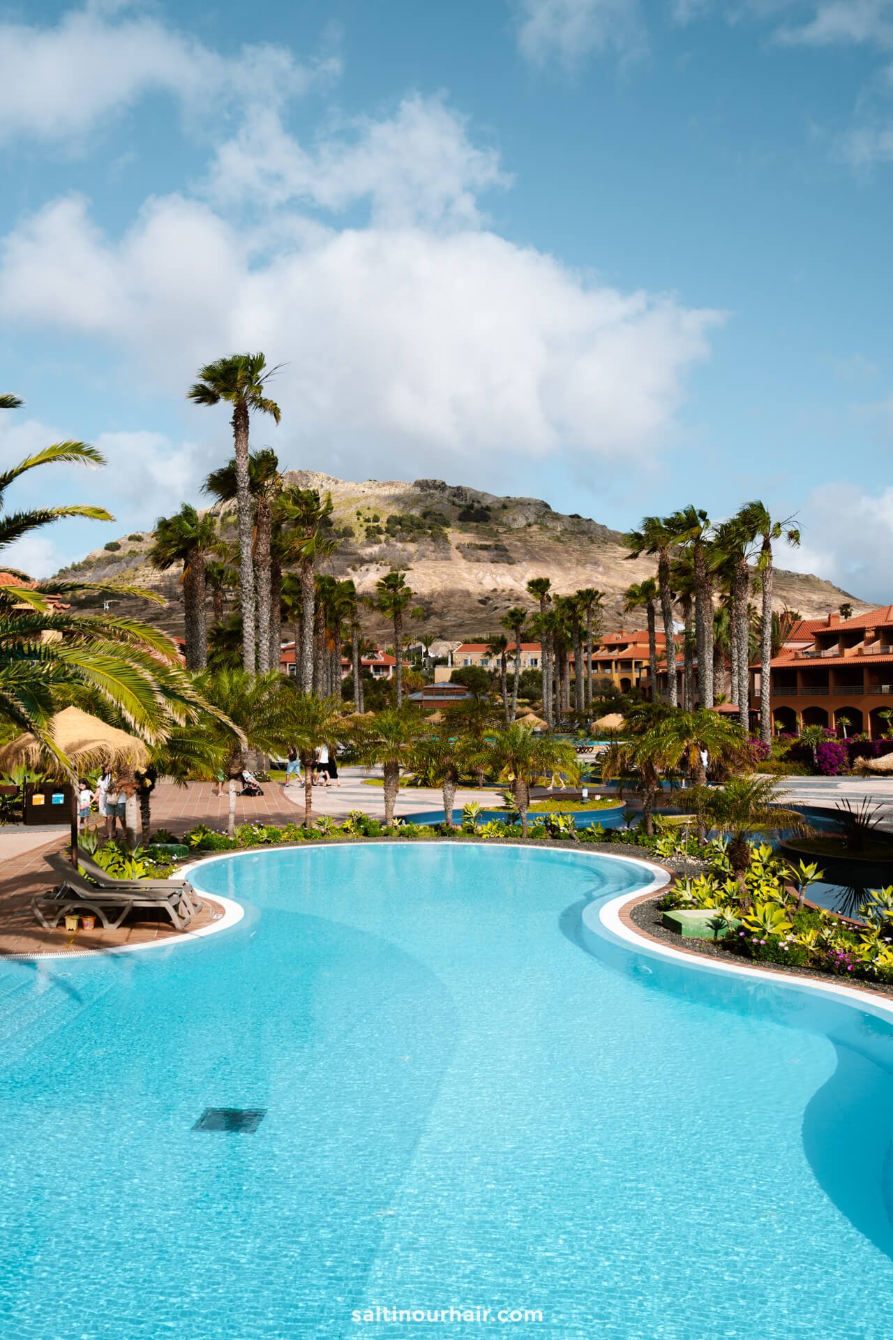 A resort pool on Porto Santo, surrounded by palm trees and tropical plants, with lounge chairs and buildings in the background, set beneath a partly cloudy sky