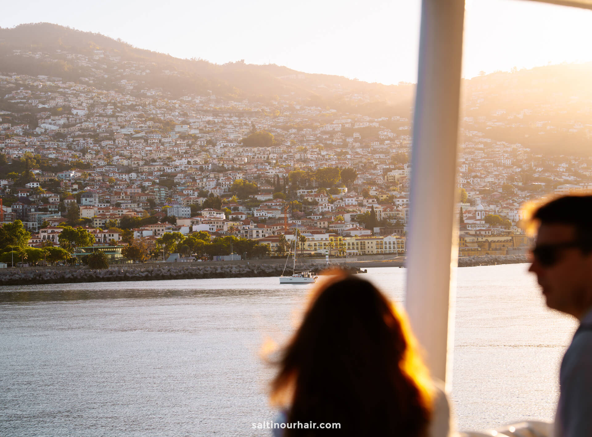 Two people stand on a waterfront deck on the ferry from Madeira to Porto Santo, overlooking a sunlit hillside town with densely packed buildings and a sailboat gliding by