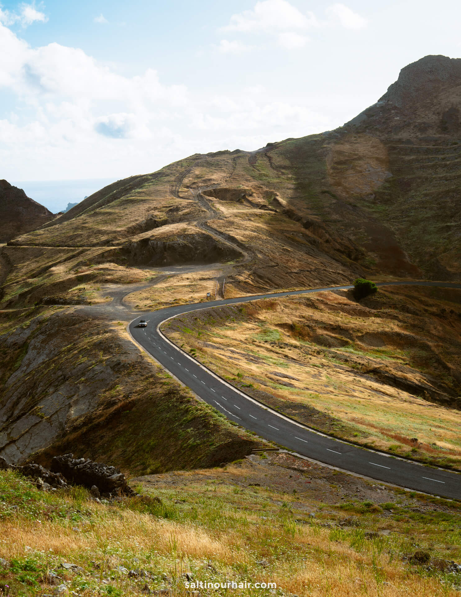 a car on a winding paved road curves through Porto Santos hilly, grassy landscape under a partly cloudy sky.