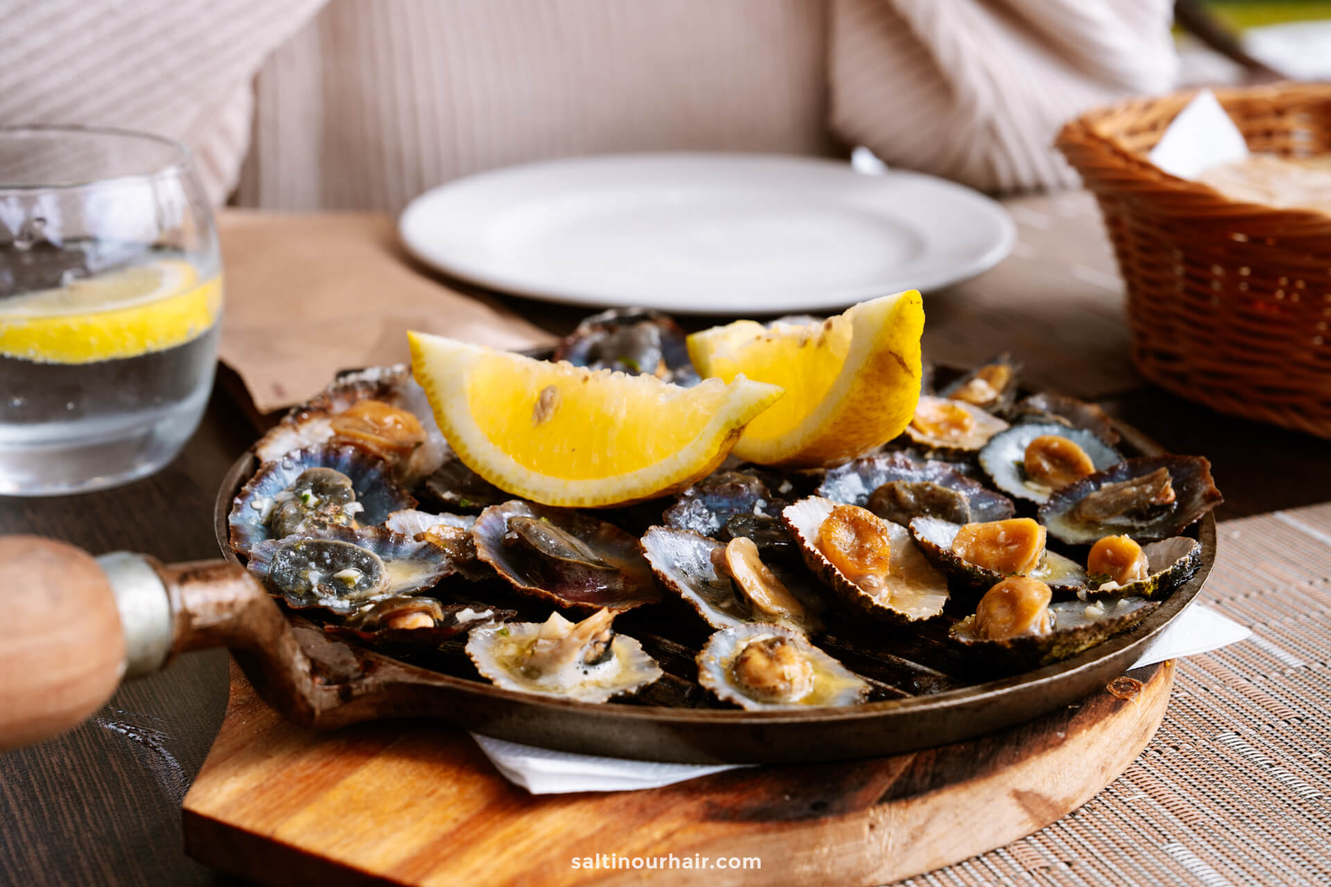 A skillet of cooked limpets with lemon wedges on top sits on a wooden board; enjoying this local dish is one of the delicious things to do in Porto Santo. A glass of water with lemon and a basket of bread are in the background.