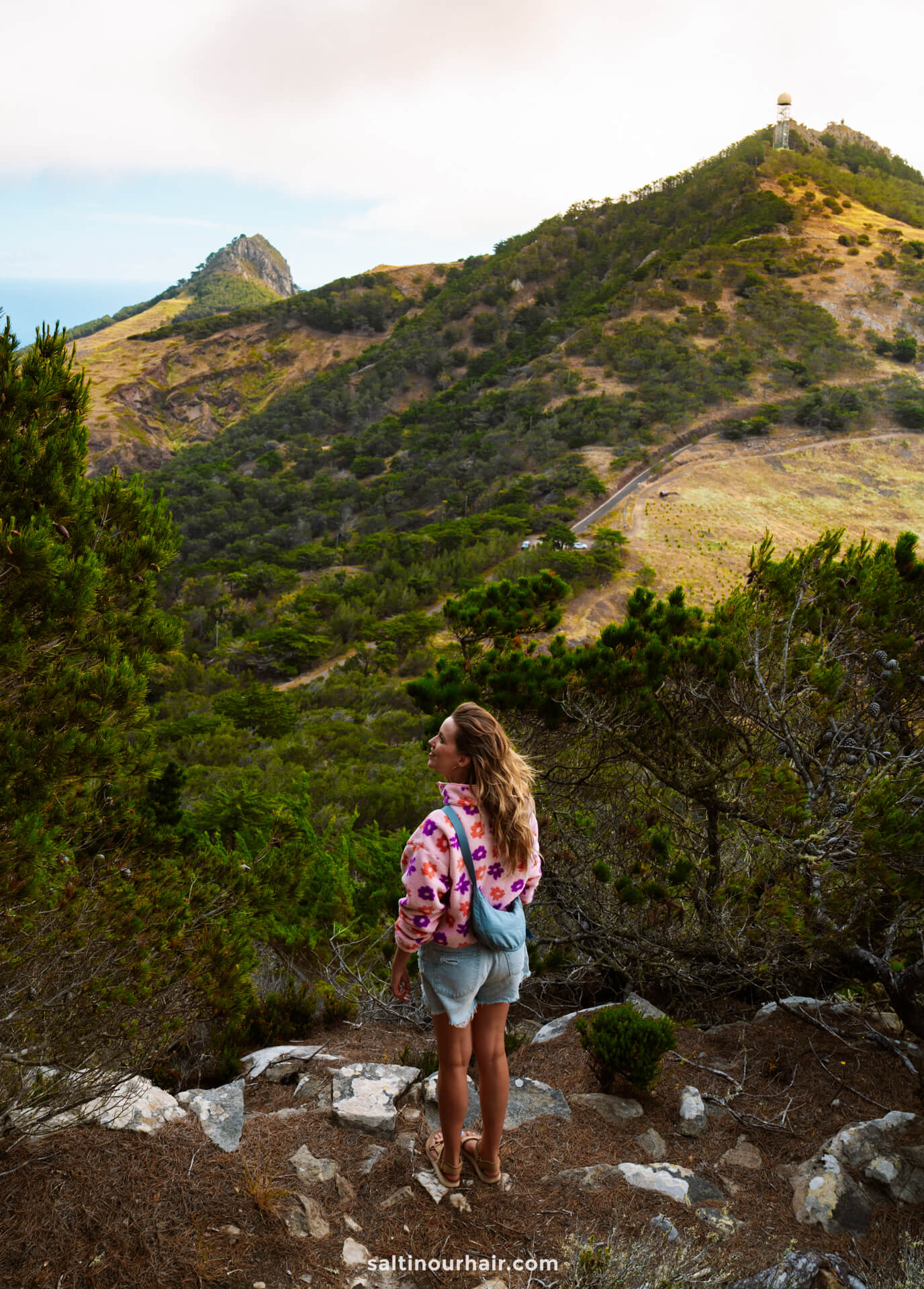 A woman in a pink and purple shirt stands on a rocky path overlooking Porto Santoâ€™s green hills and a distant hilltop structure under a cloudy sky, capturing the scenic beauty and things to do in Porto Santo.