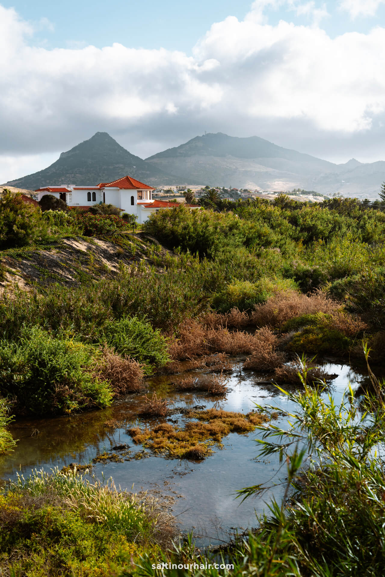 A white house with a red roof sits among green vegetation near a pond, with mountains and a cloudy sky in the backgroundâ€”capturing the peaceful charm of Porto Santo madeira