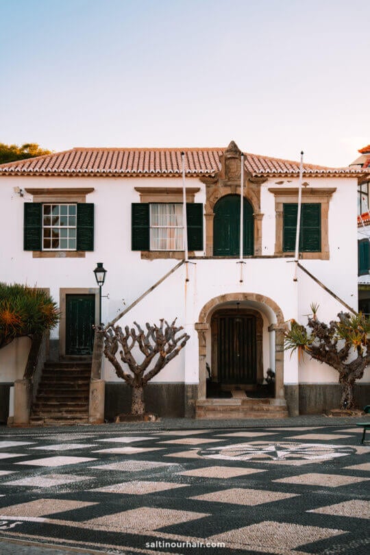 A white colonial-style building with green shutters, an arched doorway, bare trees, and a patterned stone courtyard in Porto Santo town.