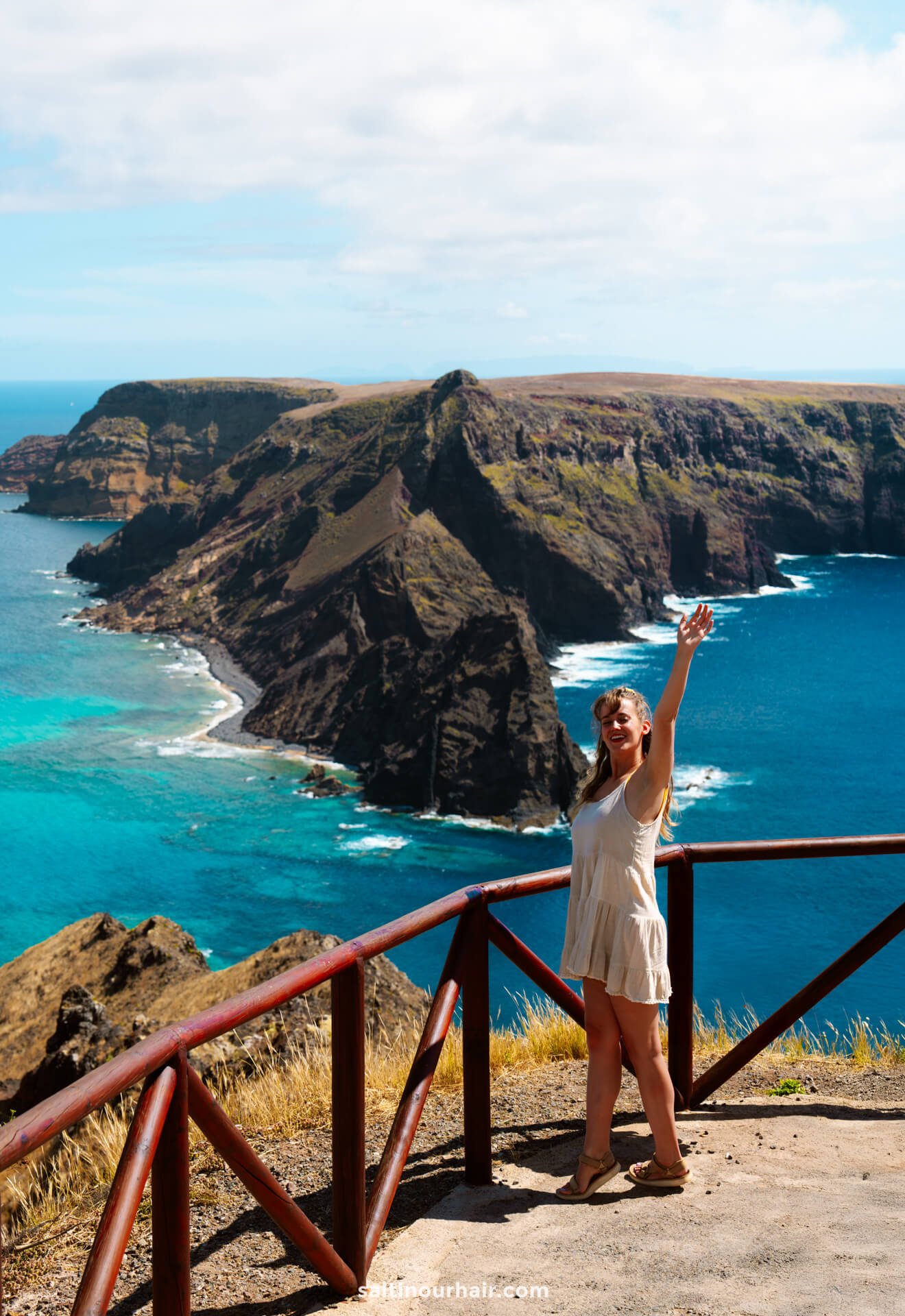 A woman stands by a wooden railing on a cliffside in Porto Santo, smiling and raising one arm, with rugged coastal cliffs and the blue ocean behind herâ€”one of the unforgettable things to do in Porto Santo.