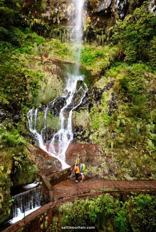 Twee wandelaars staan op een gebogen wandelpad voor een weelderige groene klif met trapsgewijze watervallen.
