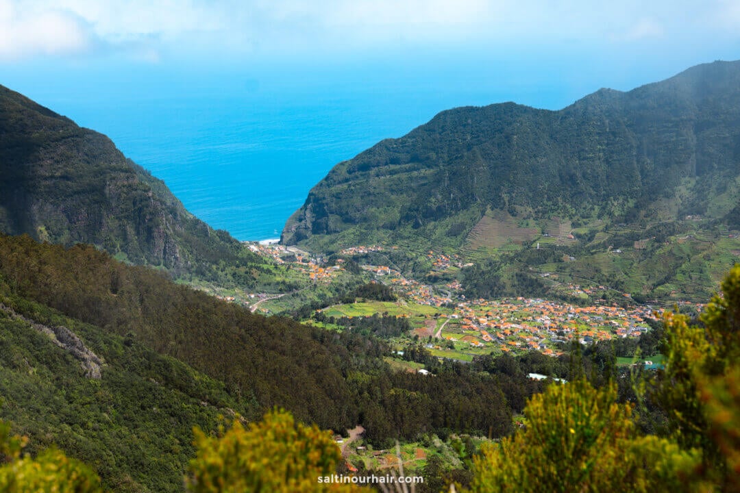 A coastal mountain valley along the PR17 Madeira route, with a small town called Sao Vicente nestled among green hills, overlooks the blue ocean under a partly cloudy sky.
