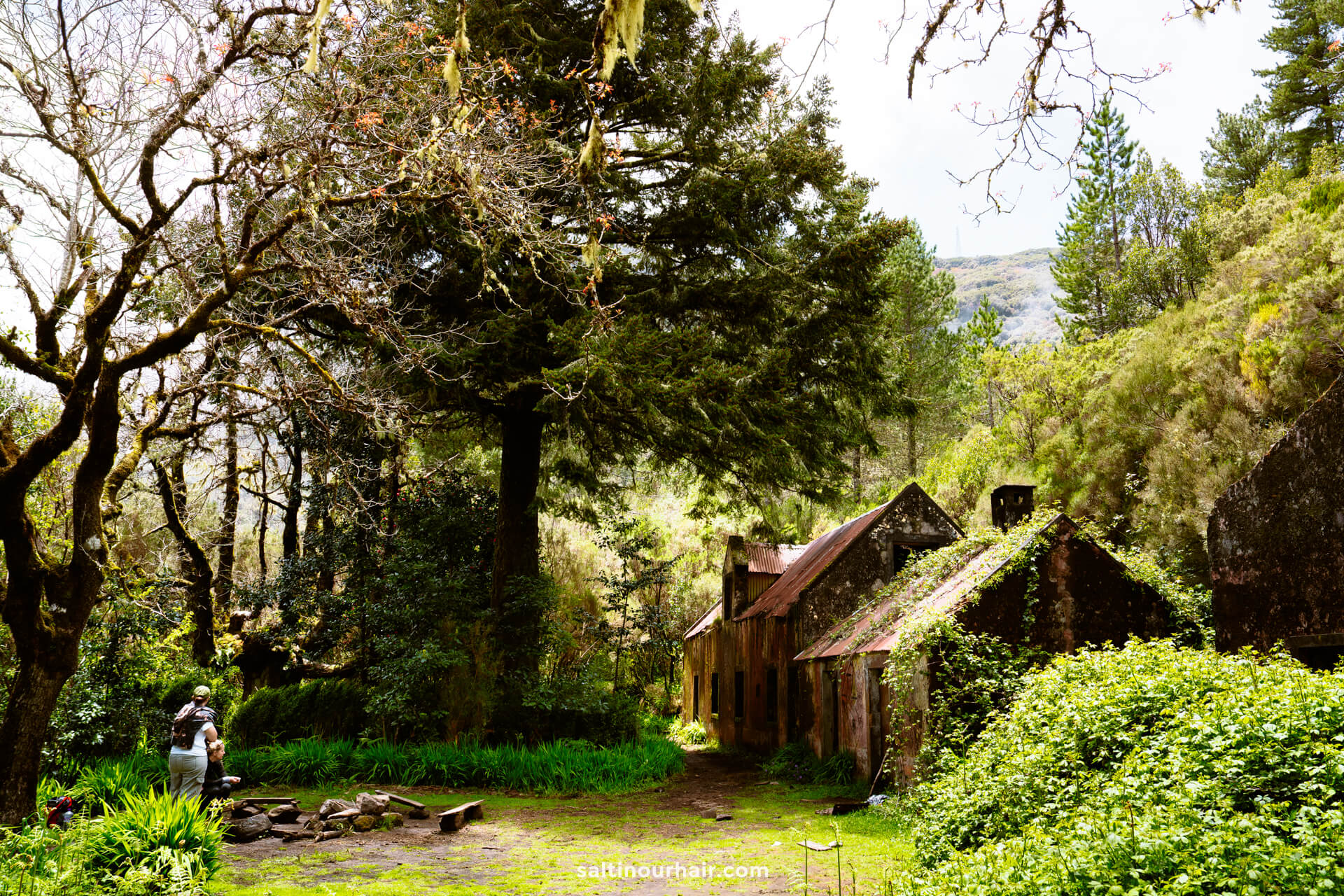 Two people stand near a stone circle along the PR17 Madeira trail in a lush, forested area with old, moss-covered buildings and tall trees surrounding them.