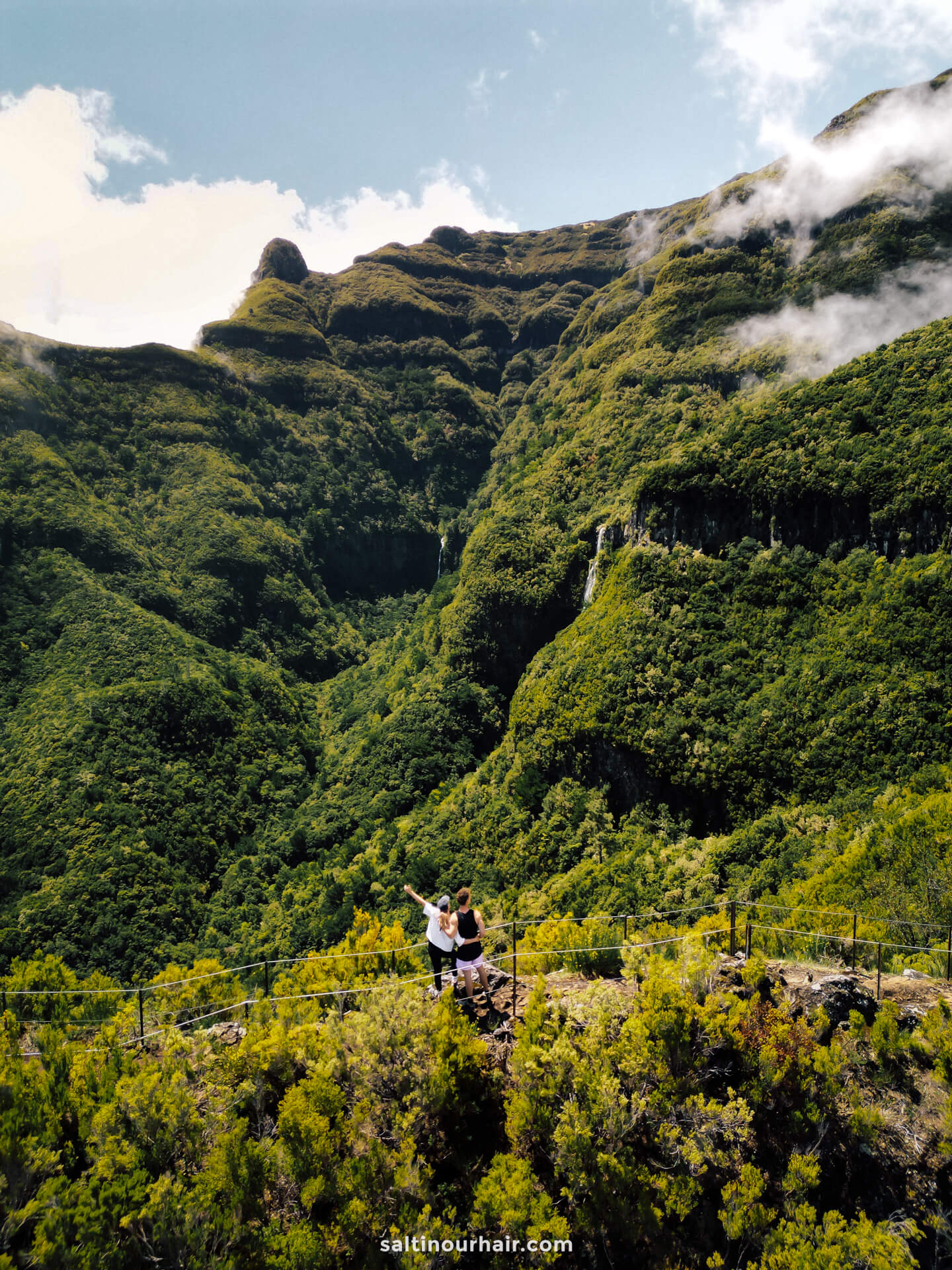 Two people stand on the PR17 Madeira trail, overlooking a lush green valley surrounded by mountains under a partly cloudy sky.