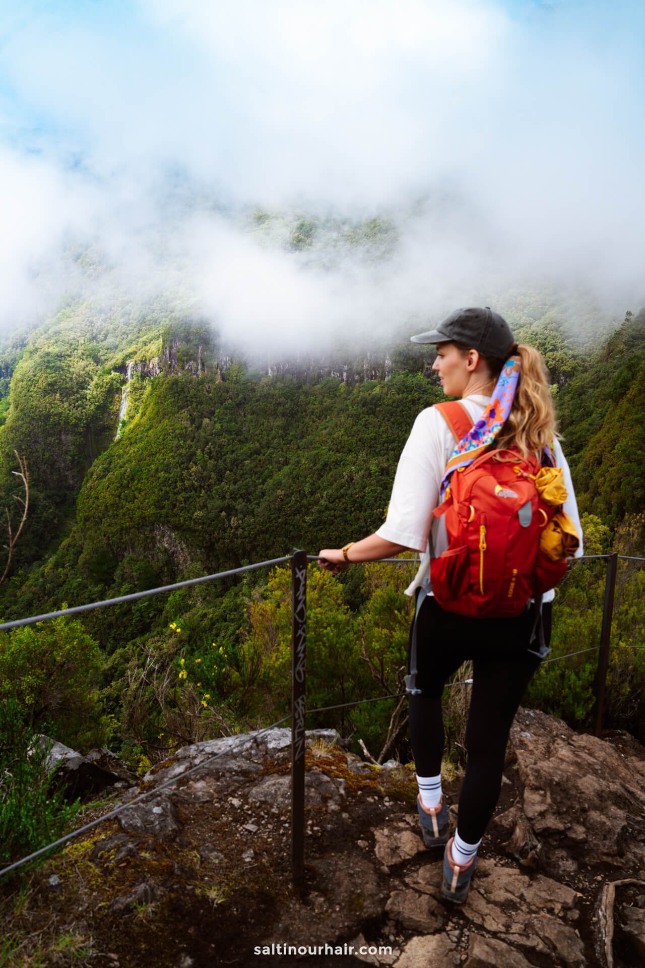 A woman with a red backpack stands on a rocky viewpoint along the PR17 Madeira trail, looking at misty green mountains partially covered by clouds.
