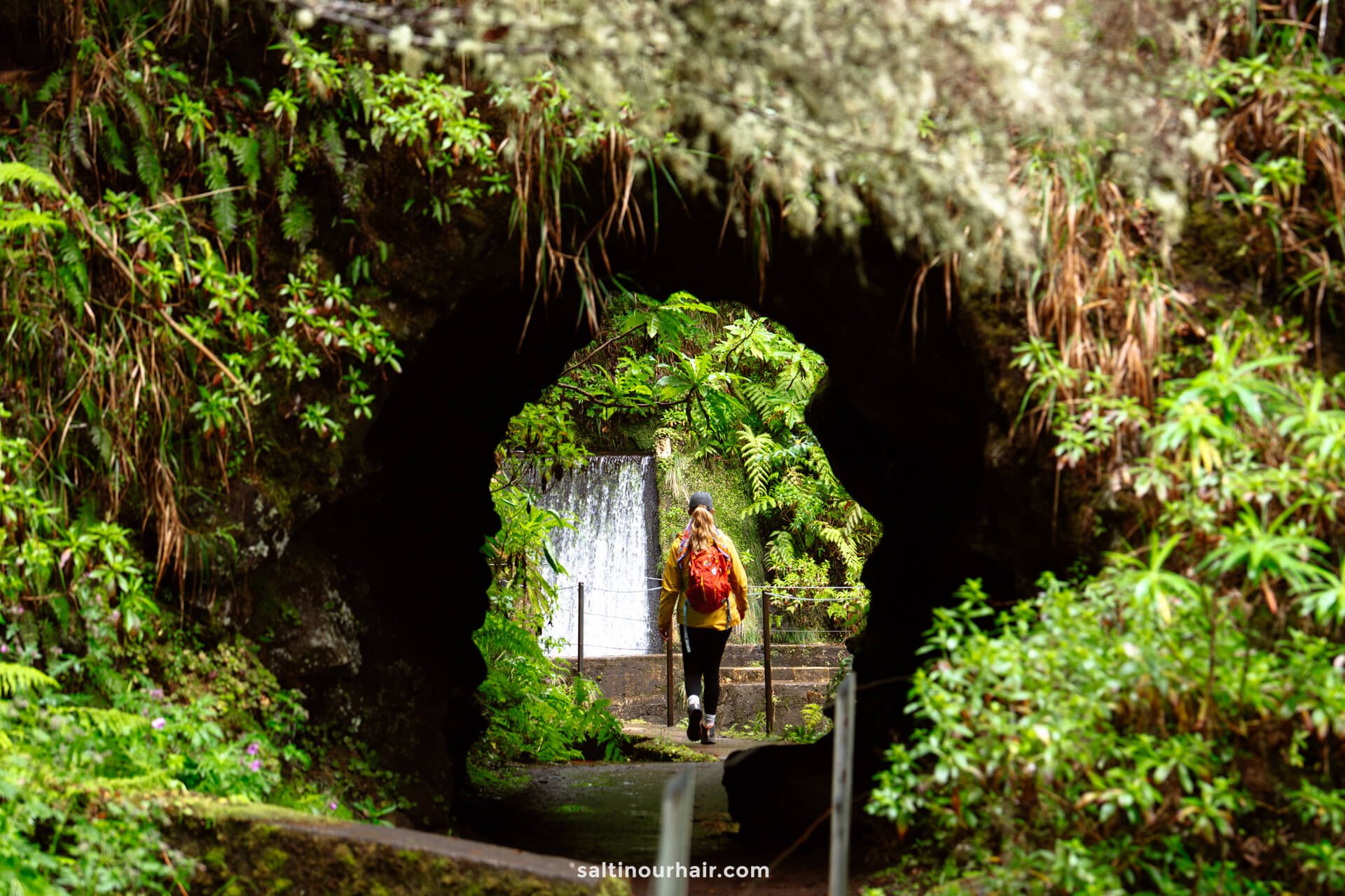 A woman with a red backpack walks through a tunnel surrounded by dense greenery along the PR17 Madeira trail, with a waterfall visible in the background.