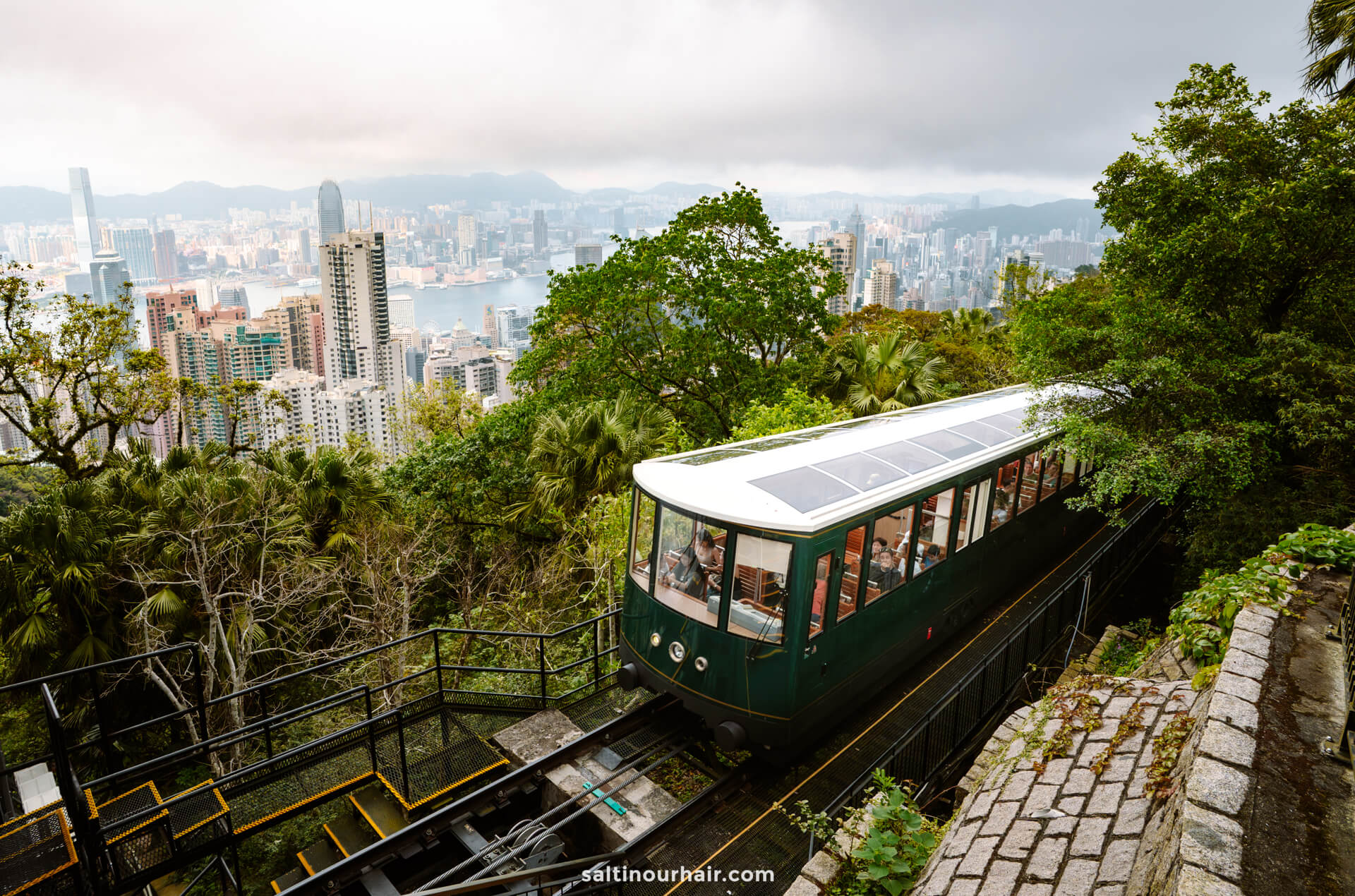 A green funicular tram ascends a steep track surrounded by trees, with a city skyline and tall buildings in the background in Hong Kong