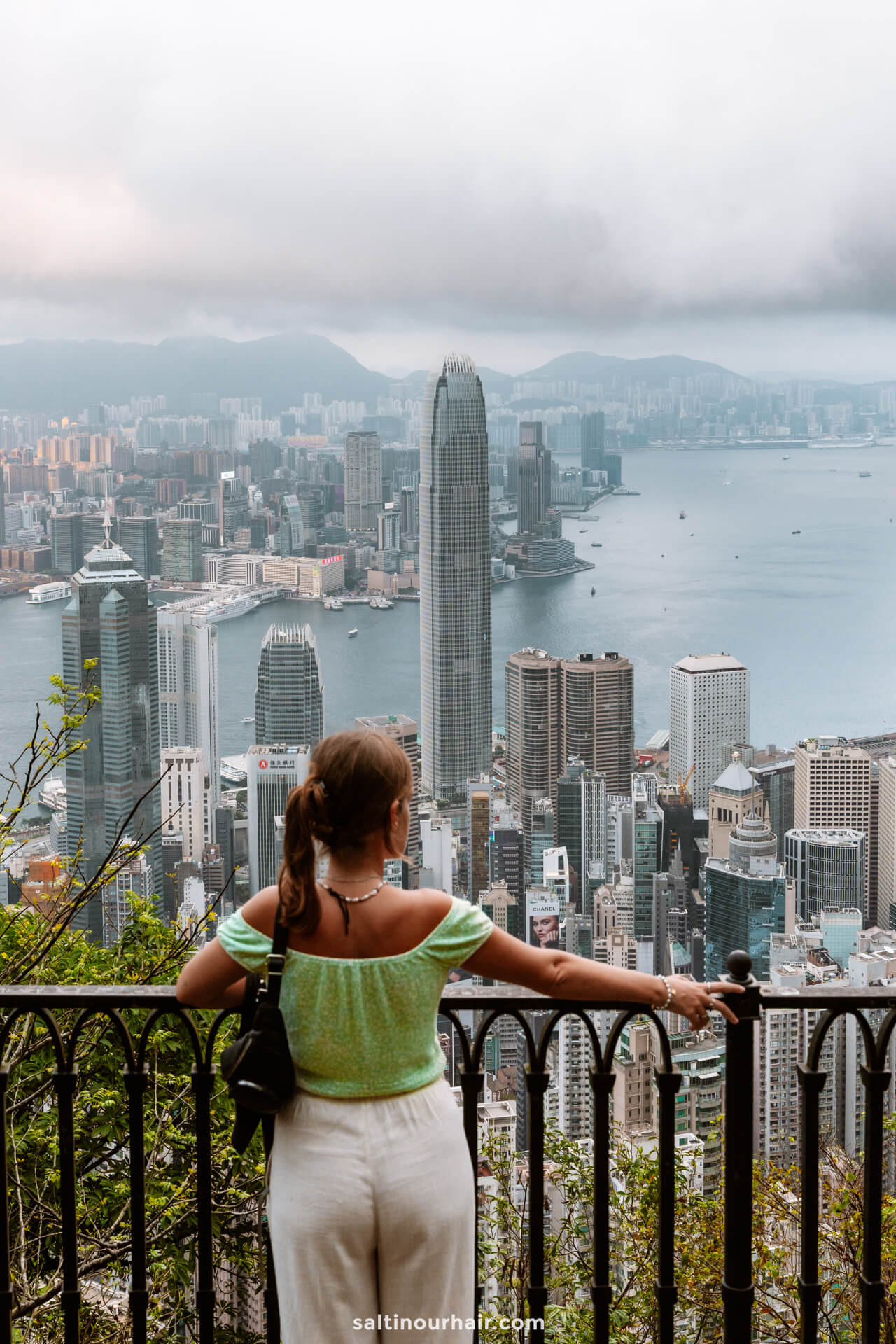 A woman stands at a railing at Victoria Peak Hong Kong overlooking the city skyline and harbor under a cloudy sky