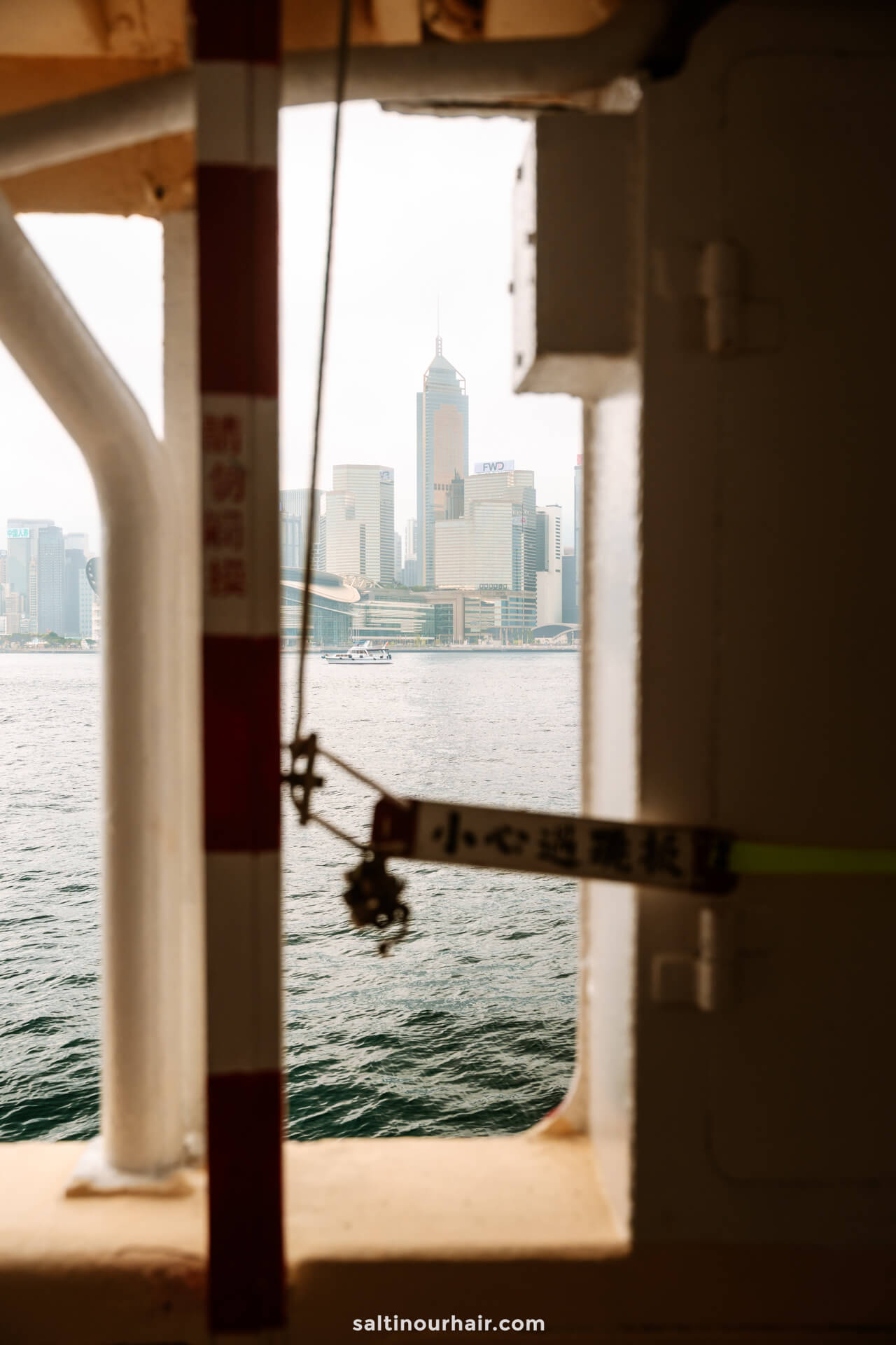 View of Hong Kong city skyline across the water, seen from a ferry with metal bars and caution tape in the foreground