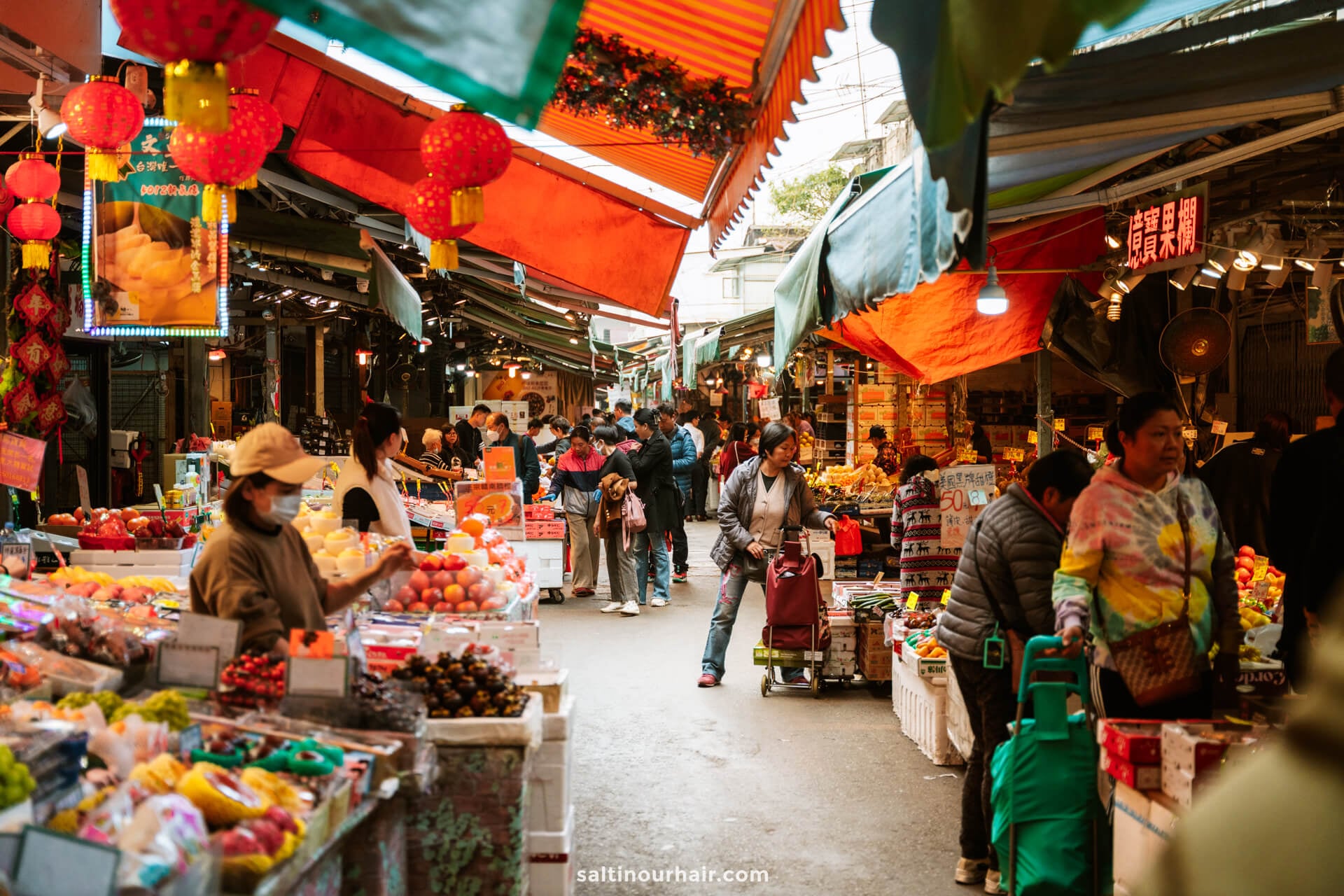 Outdoor market scene with various fruit and vegetable stalls, shoppers exploring aisles, and colorful awnings and lanterns overhead in vibrant Hong Kong
