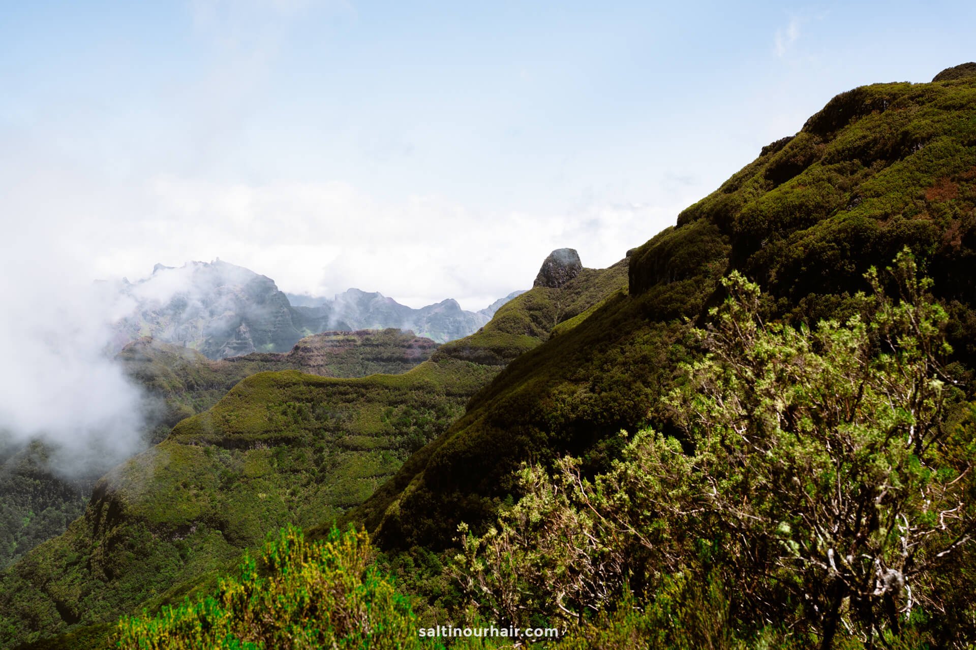 Mountain landscape along the pr17 Madeira trail, with lush green vegetation, partially covered by clouds, and distant rocky peaks under a bright, partly cloudy sky.