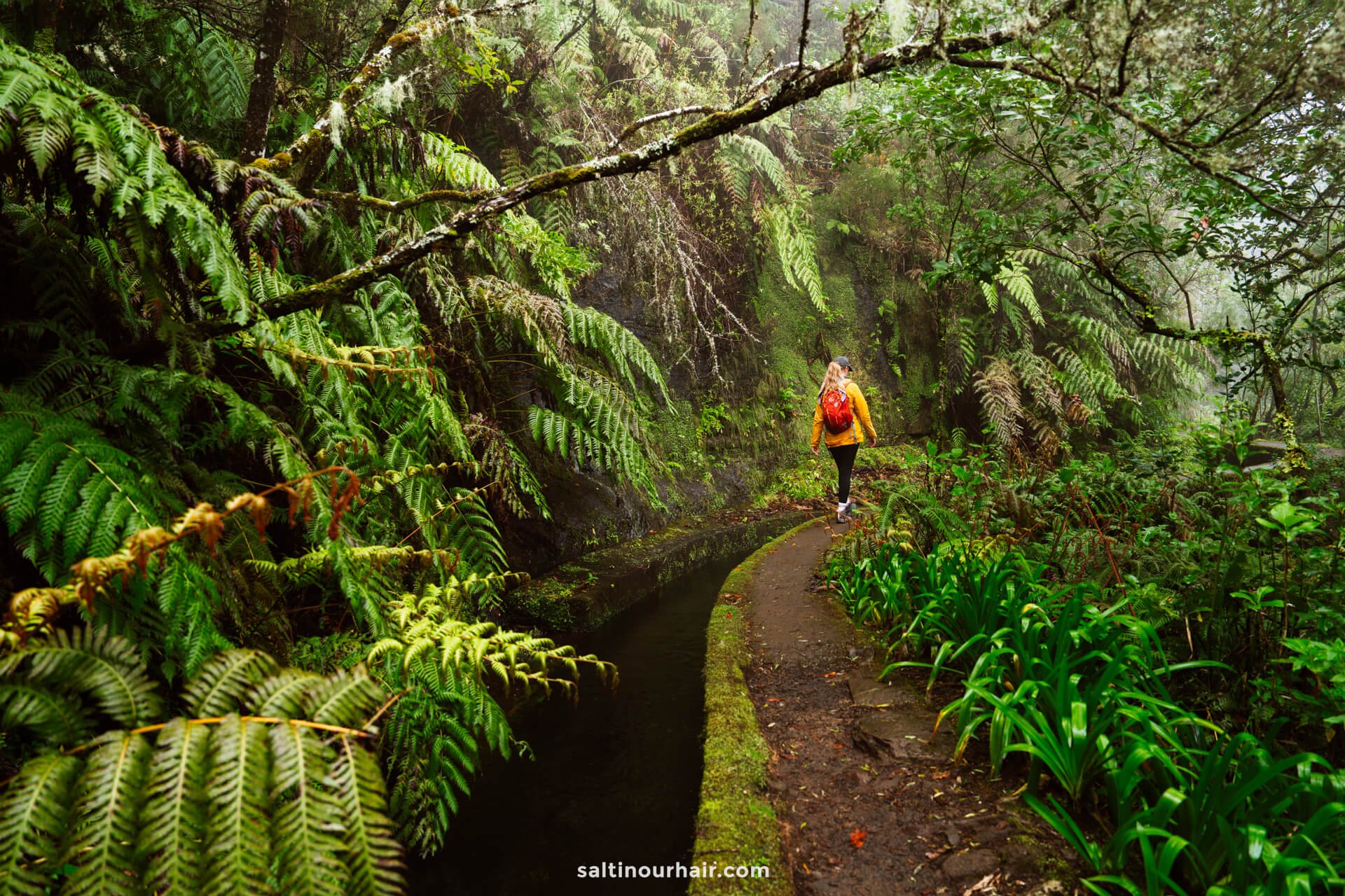 A woman with an orange backpack walks along the narrow, curved PR17 Madeira path beside a water channel in a lush, green forest.