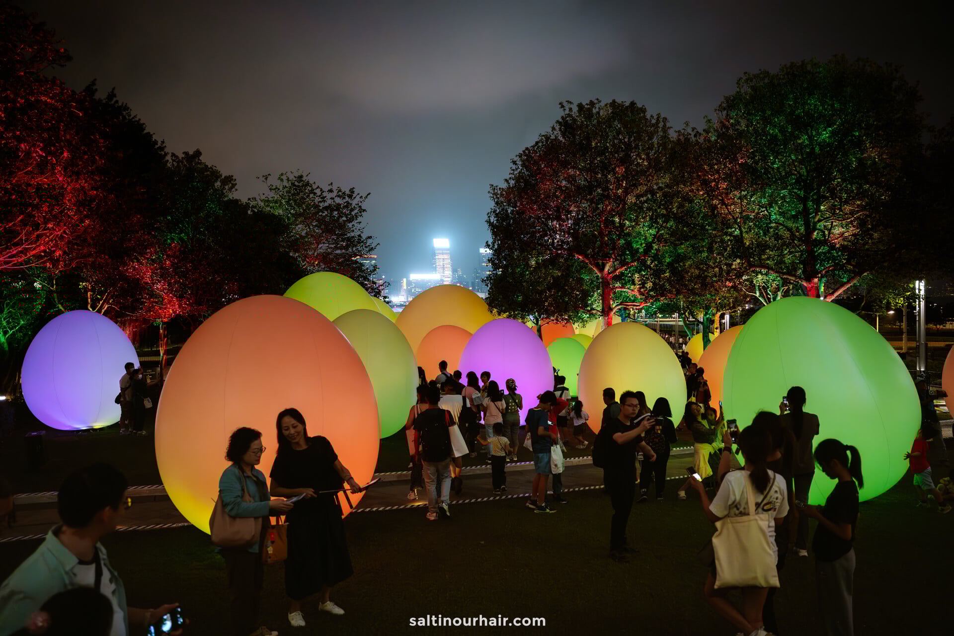 People gather in a park at night surrounded by large, illuminated, colorful egg-shaped sculptures. 