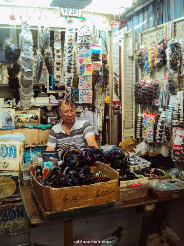 A man sits behind a counter in a shop at Causeway Bay, Hong Kong selling various hair accessories, including headbands, clips, and hair ties