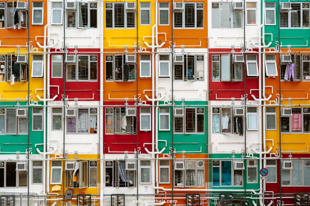 A colorful apartment building facade in Hong Kong featuring rows of windows and balconies, with red, orange, yellow, and green panels and visible pipes running vertically