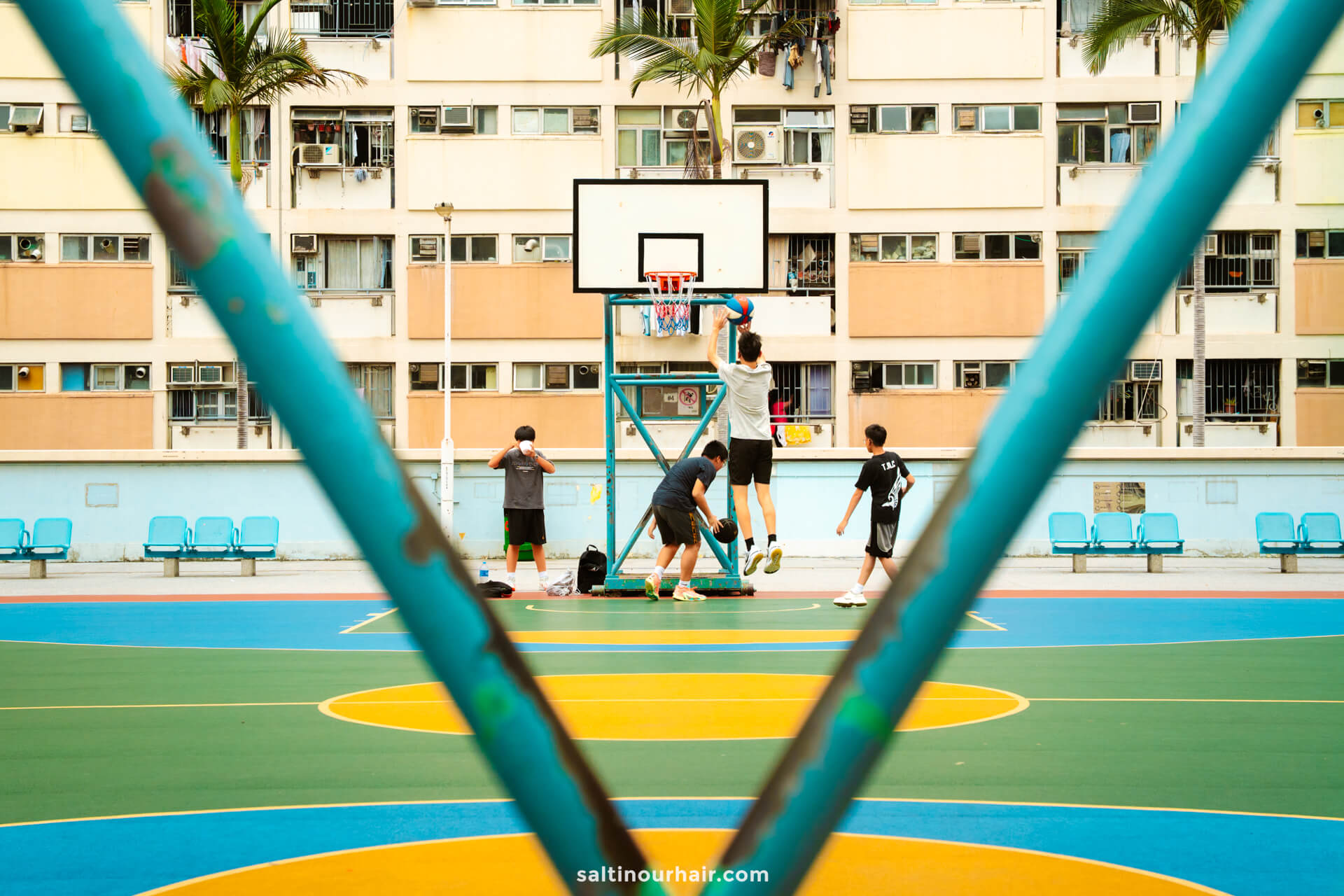 Four kids play basketball in Hong Kong on an outdoor court, framed by a blue metal structure and an apartment building