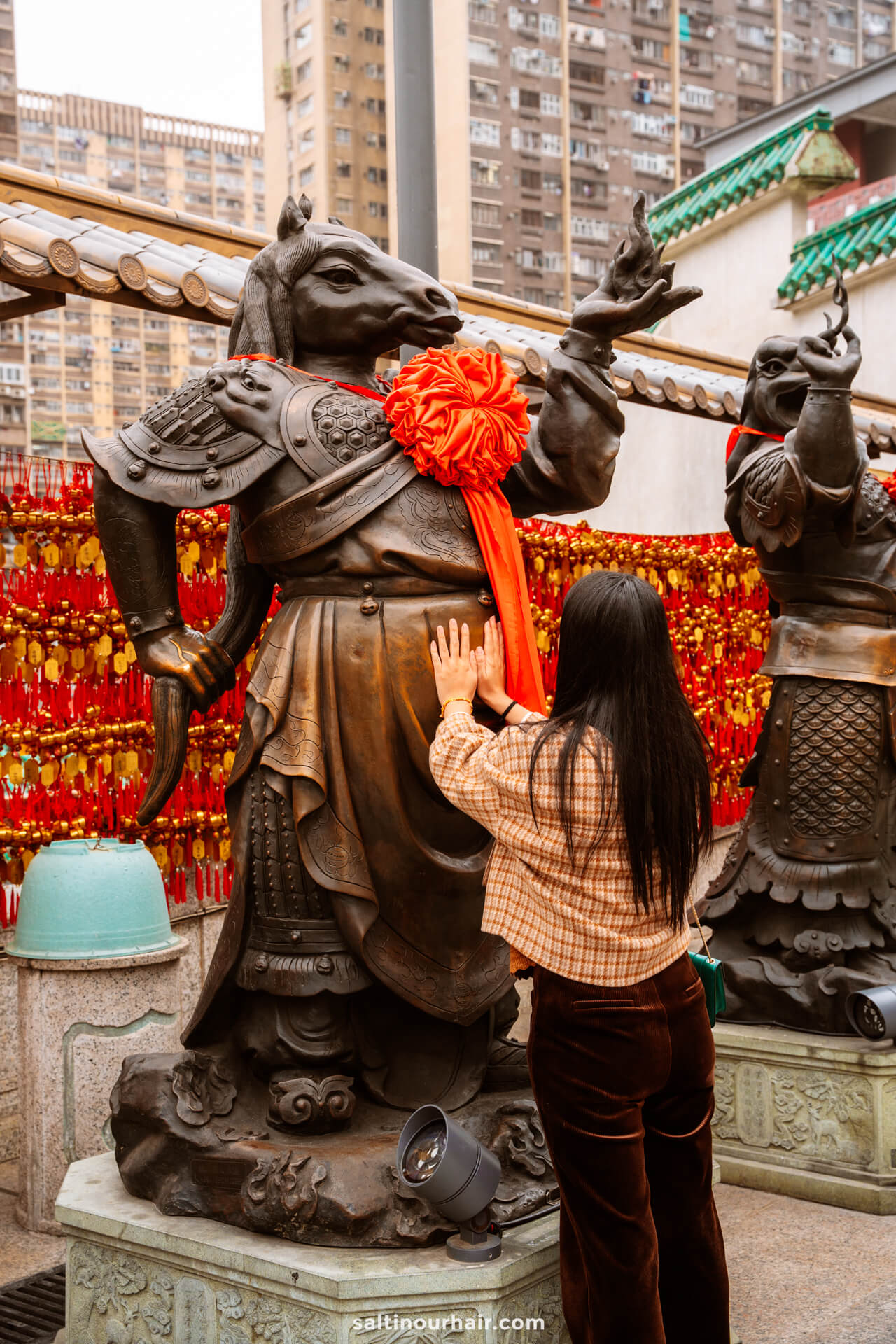 A woman touches a bronze statue of an armored animal-headed figure with a red ribbon,at Sik Sik Yuen Wong Tai Sin Temple in Hong Kong