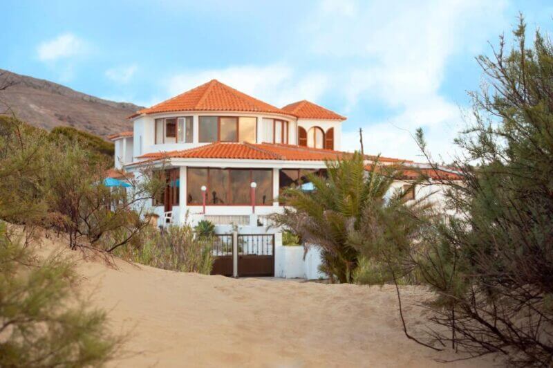 Theresia's two-story house with a red-tiled roof and large windows stands behind a gate, surrounded by Porto Santo’s sand and green shrubs.