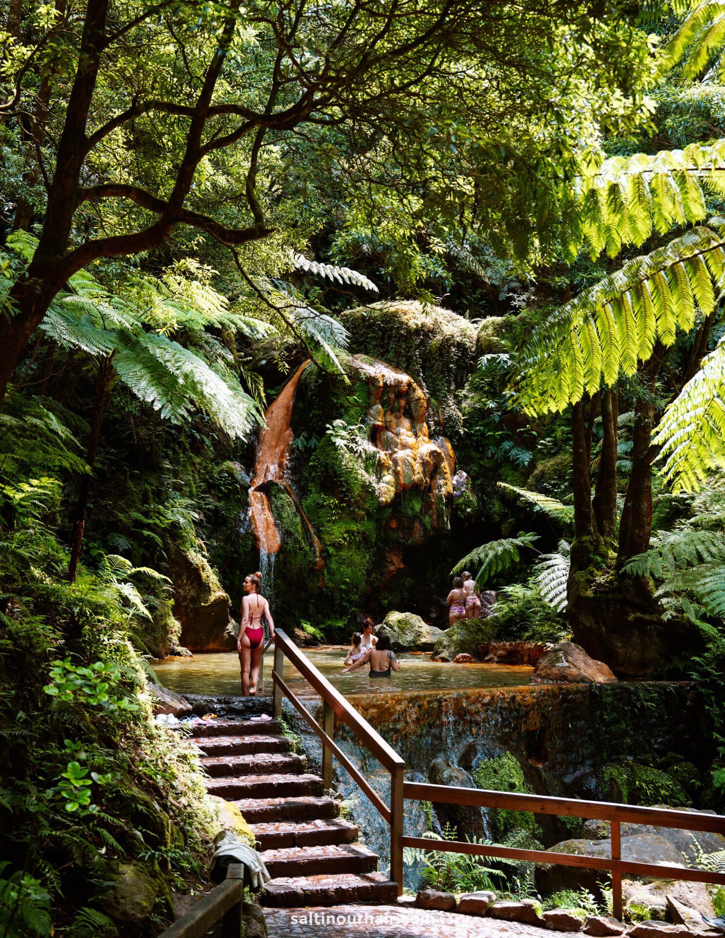 People relax in a natural hot spring pool called Caldeira Velha surrounded by lush green foliage, viewed from wooden steps leading down—an unforgettable stop on any São Miguel itinerary.
