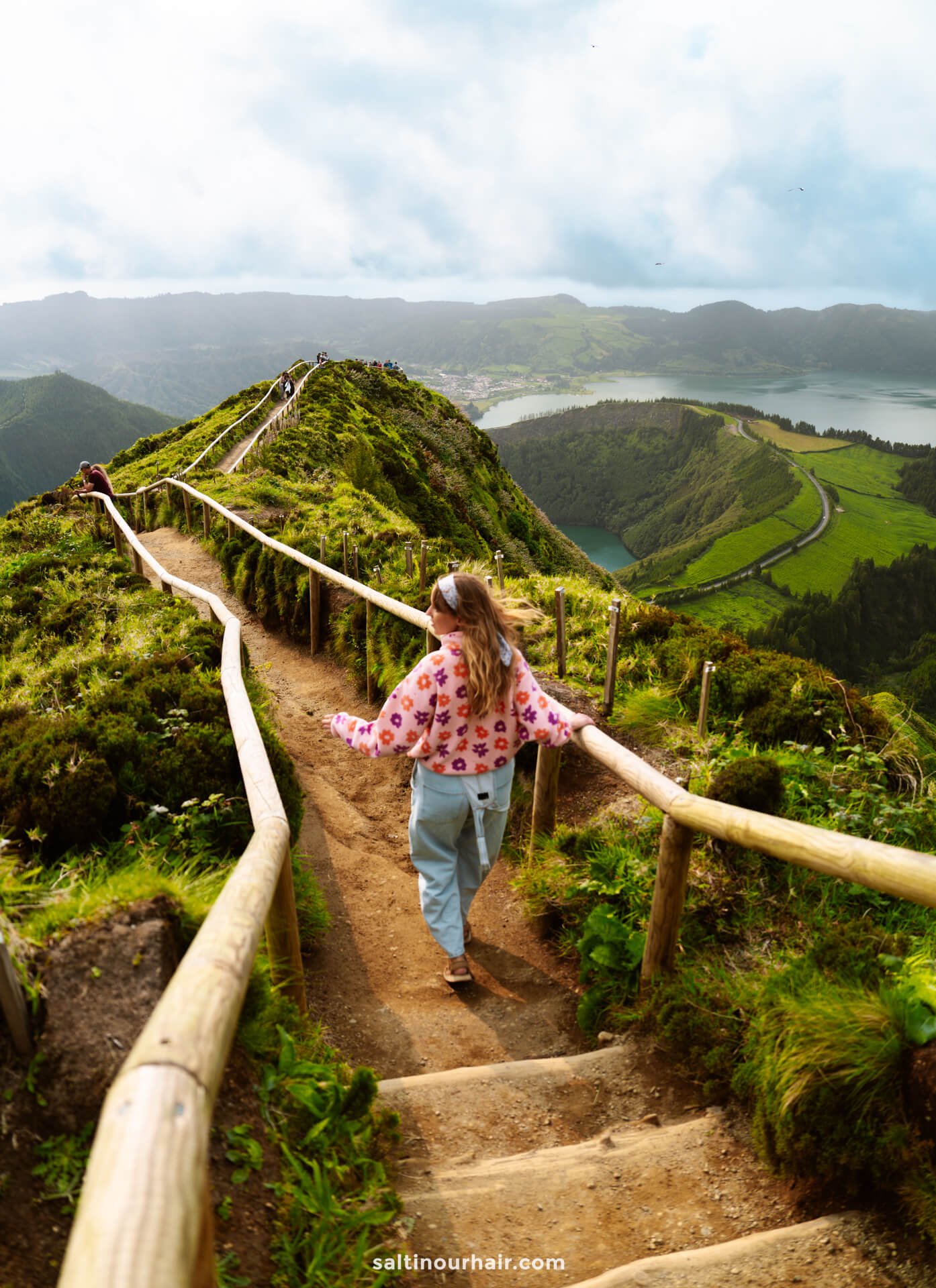 A woman walks down a dirt path at Sete Cidades Miradouro da Grota do Inferno with wooden railings along a scenic, green mountain ridge overlooking a lake and valley belowâ€”a perfect example of the breathtaking things to do in SÃ£o Miguel, Azores.