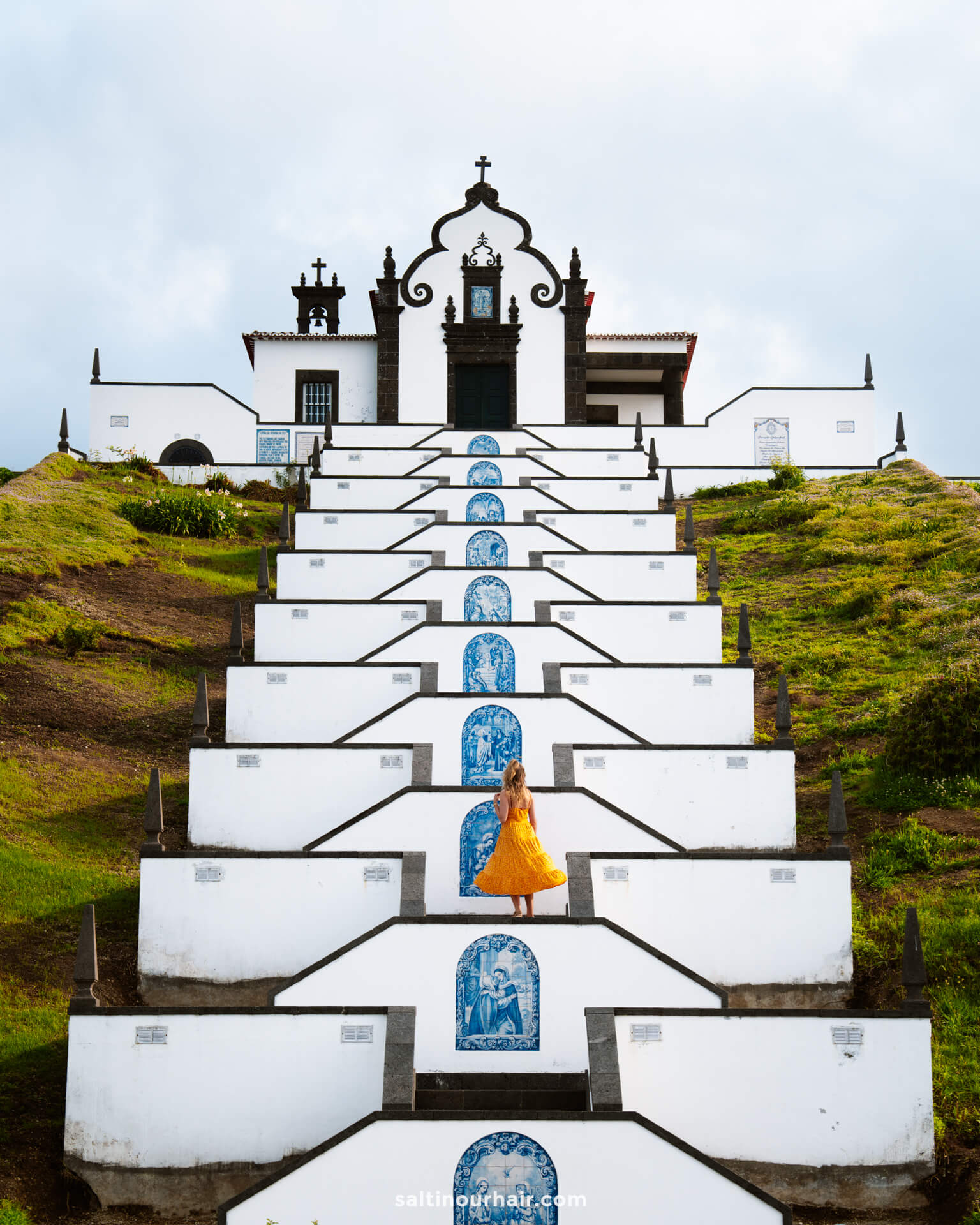 A woman in a yellow dress stands on the steps of a white church with blue tile artwork called Nossa Senhora da Paz ChapelÂ in the town of Vila Francaâ€”one of the picturesque things to do in SÃ£o Miguel, Azores.
